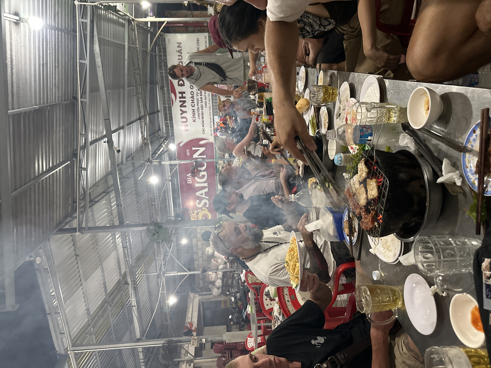 The image depicts a lively outdoor dining scene in Phu Thuy Ward, Vietnam. A large group of people is gathered around a table covered with food and drinks. A grill is in the center of the table, with someone using tongs to cook meat. Various plates, bowls, beer glasses, and water bottles are scattered across the table. Smoke from the grill fills the air. In the background, motorbikes are parked and other people are visible at nearby tables. A sign for "HUYNH Đ QUÁN" and "SAIGON" is visible. The scene is under a metal roof structure.