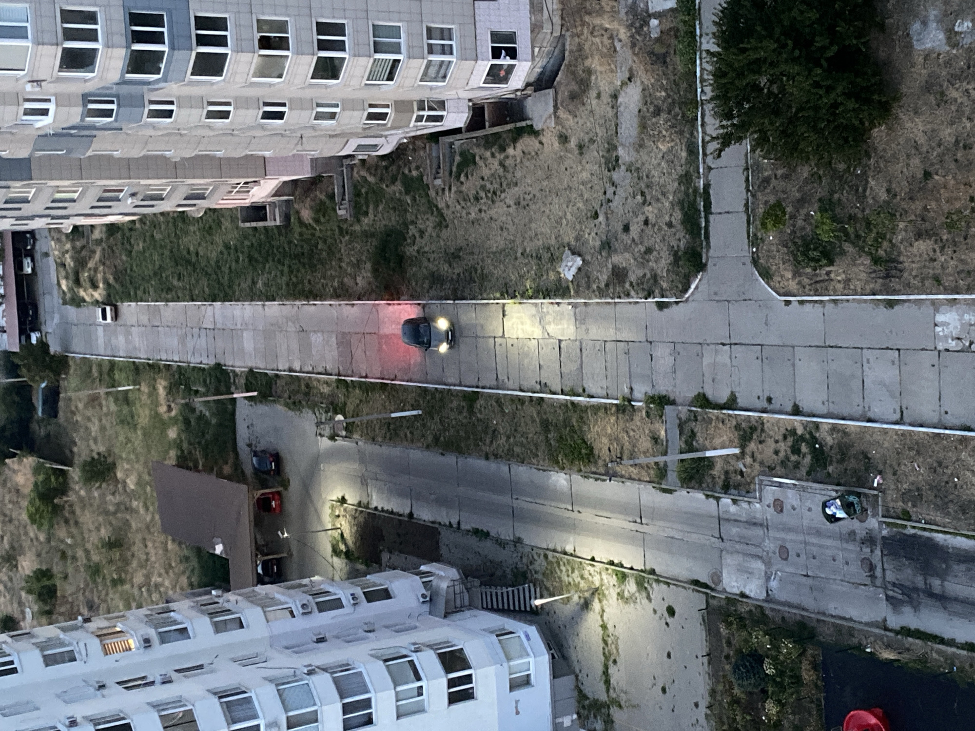 The image, taken from an elevated viewpoint, depicts a street scene in Chornomorsk, Ukraine. Two tall residential buildings dominate the left and right sides of the frame, with their multi-story facades featuring numerous windows and balconies. A paved walkway or road runs vertically through the center of the image.
On the left side of the central walkway, near the foreground, is a concrete structure containing a green trash bin overflowing with white and blue refuse. A red plastic object, possibly a playground item, is partially visible to the lower left. Adjacent to the building on the left is a set of outdoor stairs leading up to an entrance.
In the middle ground, a dark-colored car is positioned on the central walkway, with its headlights illuminated and taillights emitting a red glow. The car is facing away from the viewer. Further up the walkway, a white car is parked or driving away from the viewpoint.
To the left of the walkway, in the middle ground, a covered parking structure with a brown roof shelters three parked cars: one black, one red, and one dark-colored. The ground around the buildings and walkway is covered in sparse, dry vegetation and patches of bare earth. In the background, more dry vegetation and trees are visible. Overhead power lines with utility poles are present along the sides of the walkway. The scene appears to be an outdoor urban environment.
