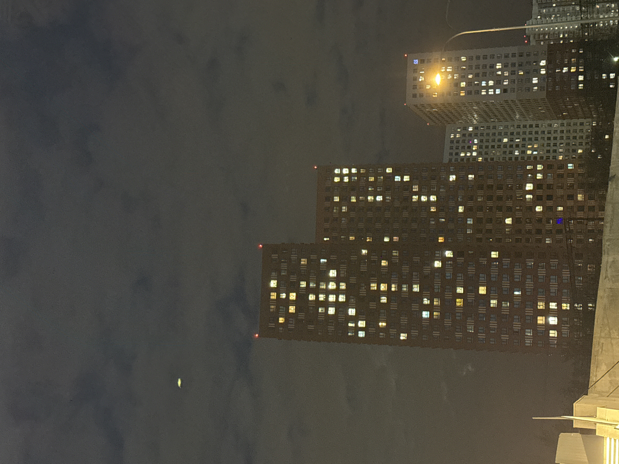 This nighttime photograph captures a dense urban scene in Khimki, Russia, dominated by several large, modern apartment or office buildings under a dark, overcast sky. The image is taken looking up at the buildings, which are vertically oriented.
In the foreground, a massive, dark-toned building with a grid-like facade and numerous illuminated rectangular windows occupies the lower and mid-right side of the frame. Small red lights, likely anti-collision beacons, are visible on its corners. Behind this structure, a lighter-colored, similar-style building stands taller, also featuring many lit windows. A bright streetlight casts a strong glow near the top of this lighter building. Further in the background, a taller, more architecturally complex building with a detailed upper section is partially visible.
The dark sky, mottled with clouds, takes up the left portion of the image. A faint, elongated, yellowish light, possibly the moon partially obscured by clouds, can be seen in the upper-left sky. On the lighter-colored building, near a bright window, a blue square logo with white letters "SS" is discernible. A concrete or stone structure with distinct edges and steps, possibly a part of an elevated walkway or another building, is partially visible on the far right. The numerous lit windows across the buildings suggest ongoing activity within the residences or offices during the nighttime.