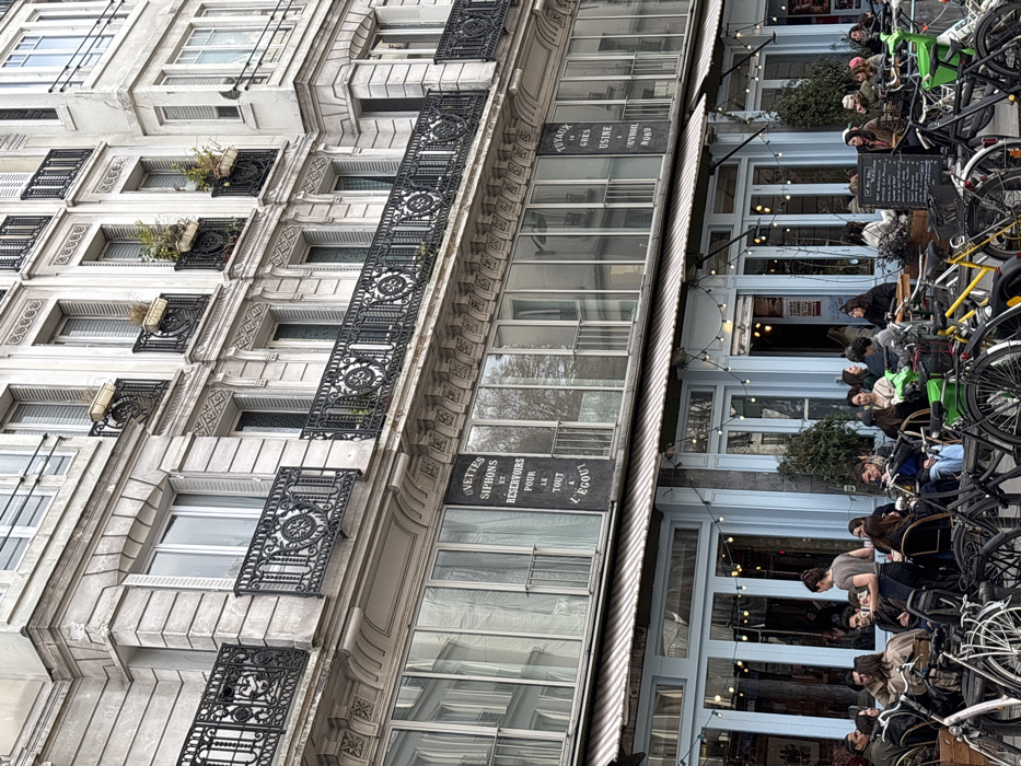 This image depicts the exterior of a multi-story Haussmannian building in Paris, France, specifically identified by historical signage as located at 92 Rue de Turenne in the 3rd arrondissement. The building features a light-colored stone facade with multiple windows, some fitted with white shutters, and ornate black wrought-iron balconies on upper floors; some balconies contain small plant pots with greenery.
Below the upper residential floors, a commercial level with large reflective glass windows displays two prominent dark signs with white lettering: the left sign reads "UVETTES SIPHONS ET RÉSERVOIRS POUR LE TOUT À L'ÉGOUT" (Valves, Siphons and Reservoirs for everything for the sewer), and the right sign reads "TUYAUX EN GRÈS USINE À CHÂTIGNOL NORD" (Stoneware pipes factory in Châtignol Nord).
The ground floor houses a cafe or restaurant with light blue-framed glass doors and windows. A striped retractable awning extends over an outdoor seating area on the sidewalk, illuminated by a string of small lights. Approximately 25-30 individuals are seated at circular wooden tables and chairs, engaged in conversations, dining, or drinking. One female individual, wearing a grey top and dark apron, stands centrally, appearing to serve patrons. In the foreground, numerous bicycles, including identifiable yellow and green rental bikes, are parked, partially obscuring the lower portion of the seated individuals. A blackboard sign displaying a menu is visible on the right side of the outdoor seating area. Small trees or shrubs in planter boxes are placed around the perimeter of the outdoor dining space. The sky appears overcast.