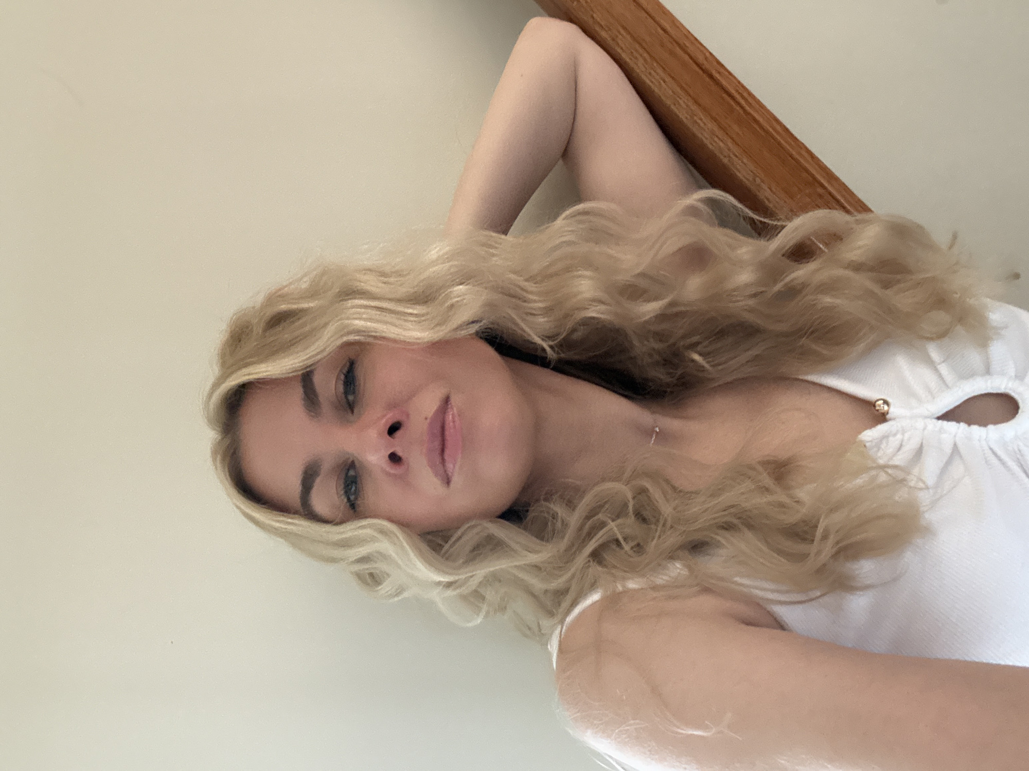 A woman with blonde, wavy hair is leaning on a wooden handrail on a staircase. She is wearing a white top with a low neckline and a delicate necklace. Her right arm is bent, and she is resting her head on her arm. The background is a plain, light-colored wall. The lighting appears to be natural.