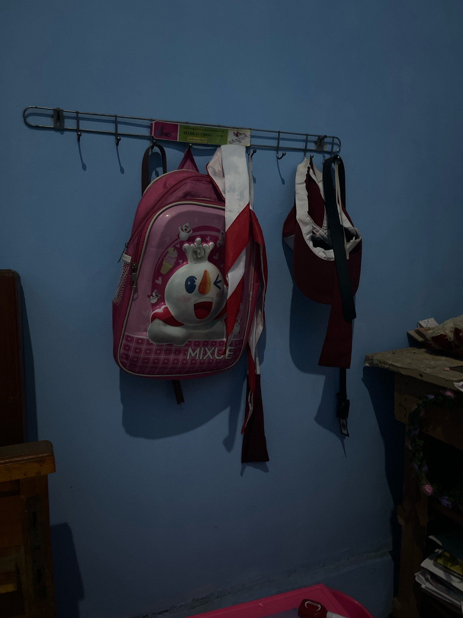 This is a medium shot, eye-level, outdoor, daylight photograph of a blue wall with a metal coat rack attached, holding two backpacks and a scarf. The backpack on the left is pink with a white cartoon character and the text "MIXUE" on it. The backpack on the right is dark red with white trim. A red and white striped scarf hangs from the coat rack between the two backpacks. To the left of the coat rack is the edge of a wooden chair or table. To the right of the coat rack is a shelf with some papers and decorative items. The overall scene suggests a home environment, possibly in the city of Kebumen, Indonesia.