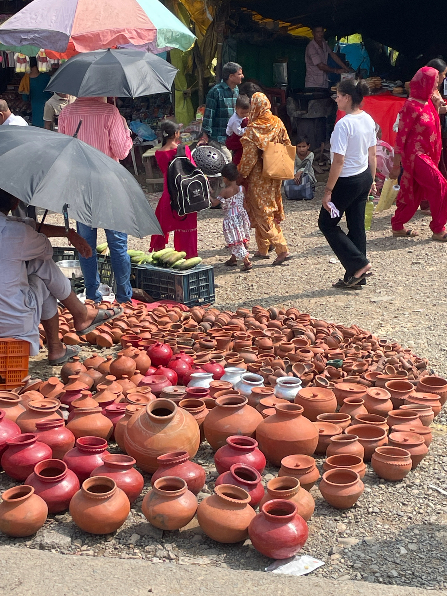The image depicts an outdoor market scene, likely in Baud, India, characterized by a bustling atmosphere and numerous vendors. The foreground is dominated by a vast collection of terracotta pottery, ranging in size and shape from small bowls to large urns, laid out on the ground. Several people are present, including women in traditional attire and men, some of whom appear to be customers or vendors. Umbrellas, both open and closed, are visible, suggesting protection from the sun or possibly rain. In the background, market stalls and awnings are set up, with more people and various goods being sold. The ground is a mixture of dirt and gravel. The overall impression is one of a lively, everyday marketplace.