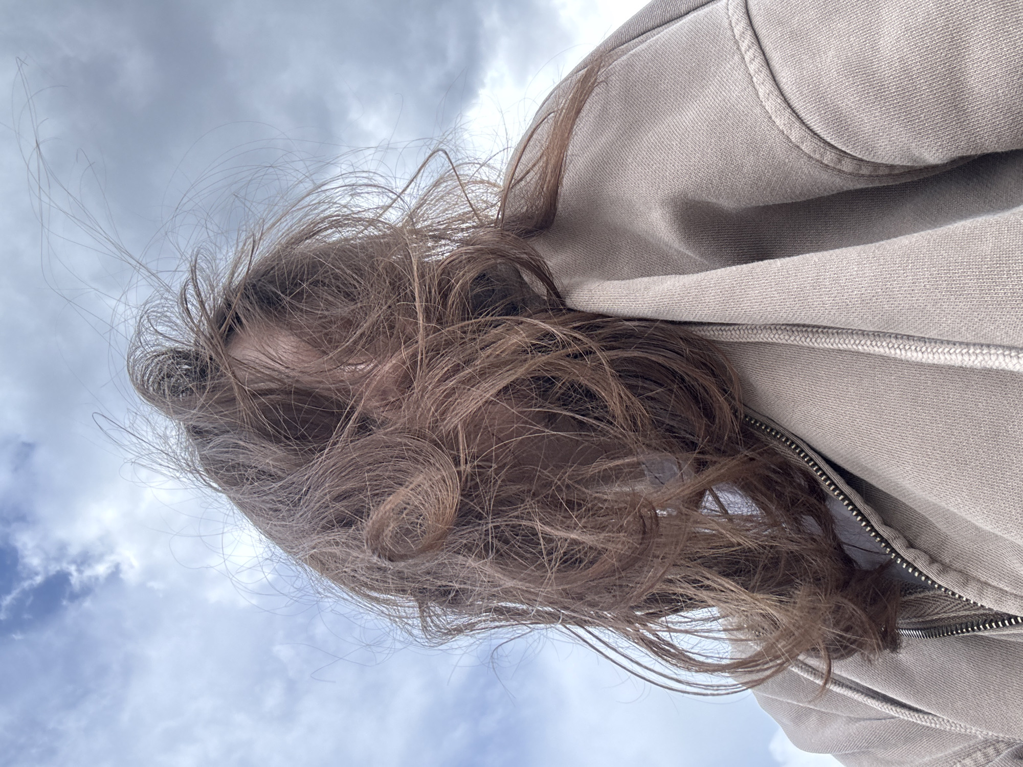 The image shows a close-up, low-angle shot of a person's head and shoulders against a cloudy sky. The person's long, brown hair is windswept and partially obscures their face. They are wearing a light-colored hooded sweatshirt with a zipper. The background consists of a dynamic sky with both light and dark clouds, suggesting movement or an impending change in weather. There is no discernible location in the city of Kazan, Russia.