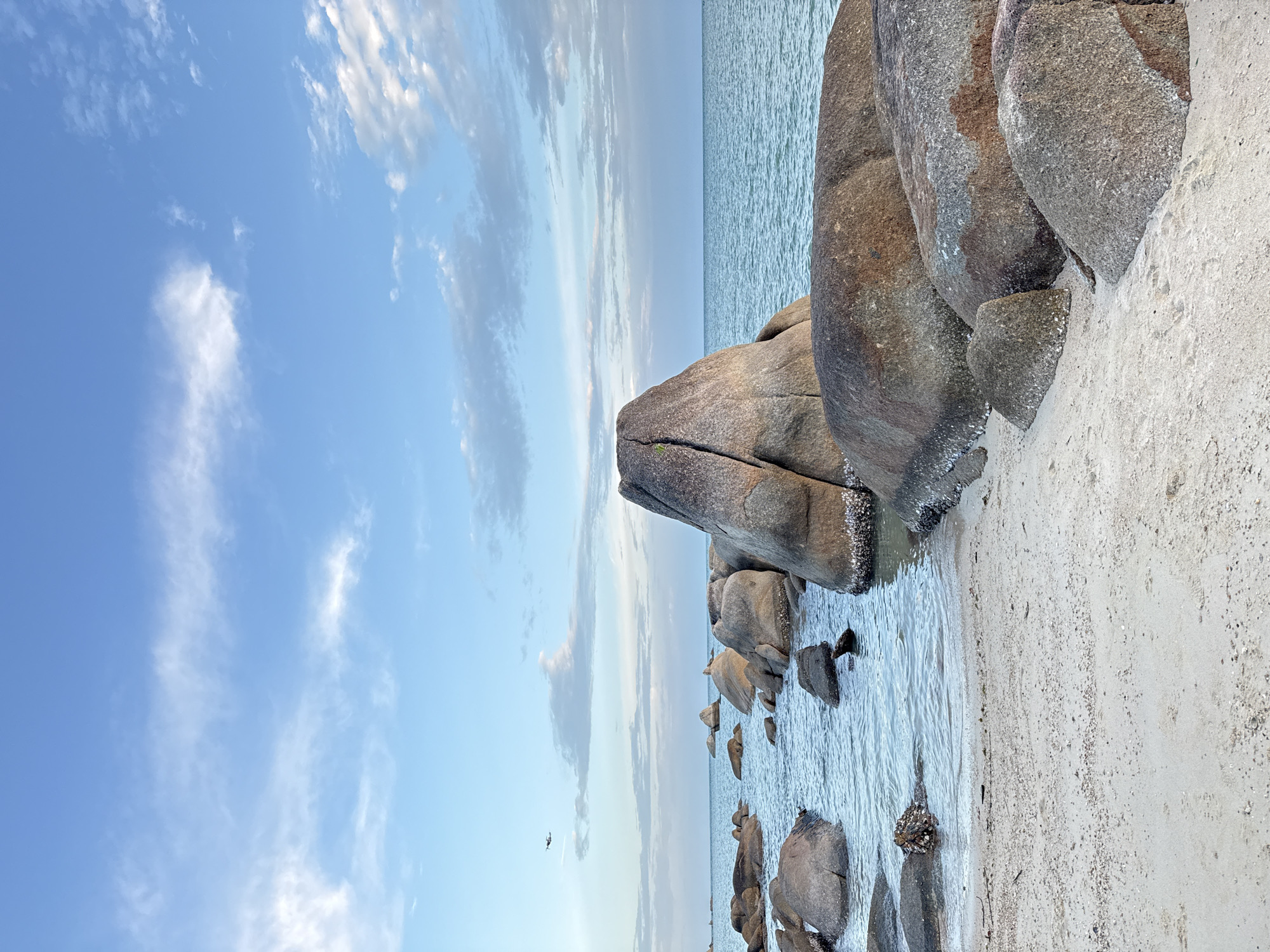 The image shows a beach scene in the Ko Samui District of Thailand. The foreground features a sandy beach with a cluster of large, weathered rocks. Some of these rocks are partially submerged in the calm, blue sea. The water is clear, and small waves are lapping at the shore. In the distance, the horizon is marked by the meeting of the sea and a pale blue sky. The sky is dotted with scattered white clouds, some of which are wispy and elongated. In the upper left quadrant of the image, a small, light-colored aircraft is visible flying in the distance. There are no people or animals clearly depicted in the image. The overall impression is one of a serene, natural coastal landscape.