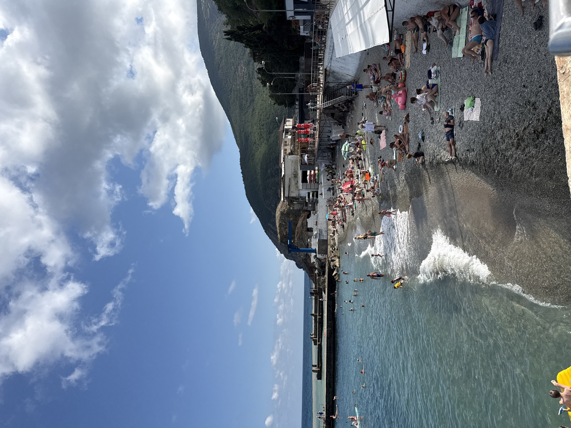 The image depicts a lively beach scene in Partenit, Ukraine, on a bright, partly cloudy day. A rocky shore is dotted with numerous people sunbathing and relaxing on towels and mats. A gentle wave washes ashore, with several individuals wading or swimming in the clear, turquoise water.
In the background, a concrete pier juts out into the sea, with a few people visible on it. To the right of the beach, a hillside covered in lush green vegetation rises dramatically. Several buildings, including what appears to be a hotel or resort complex with red umbrellas, are nestled against the hillside. A blue construction crane is prominently positioned near the water's edge.
The sky is a vibrant blue, with scattered white cumulus clouds adding texture. The overall atmosphere is one of a popular tourist destination, with people enjoying a day by the sea under the warm sun.
