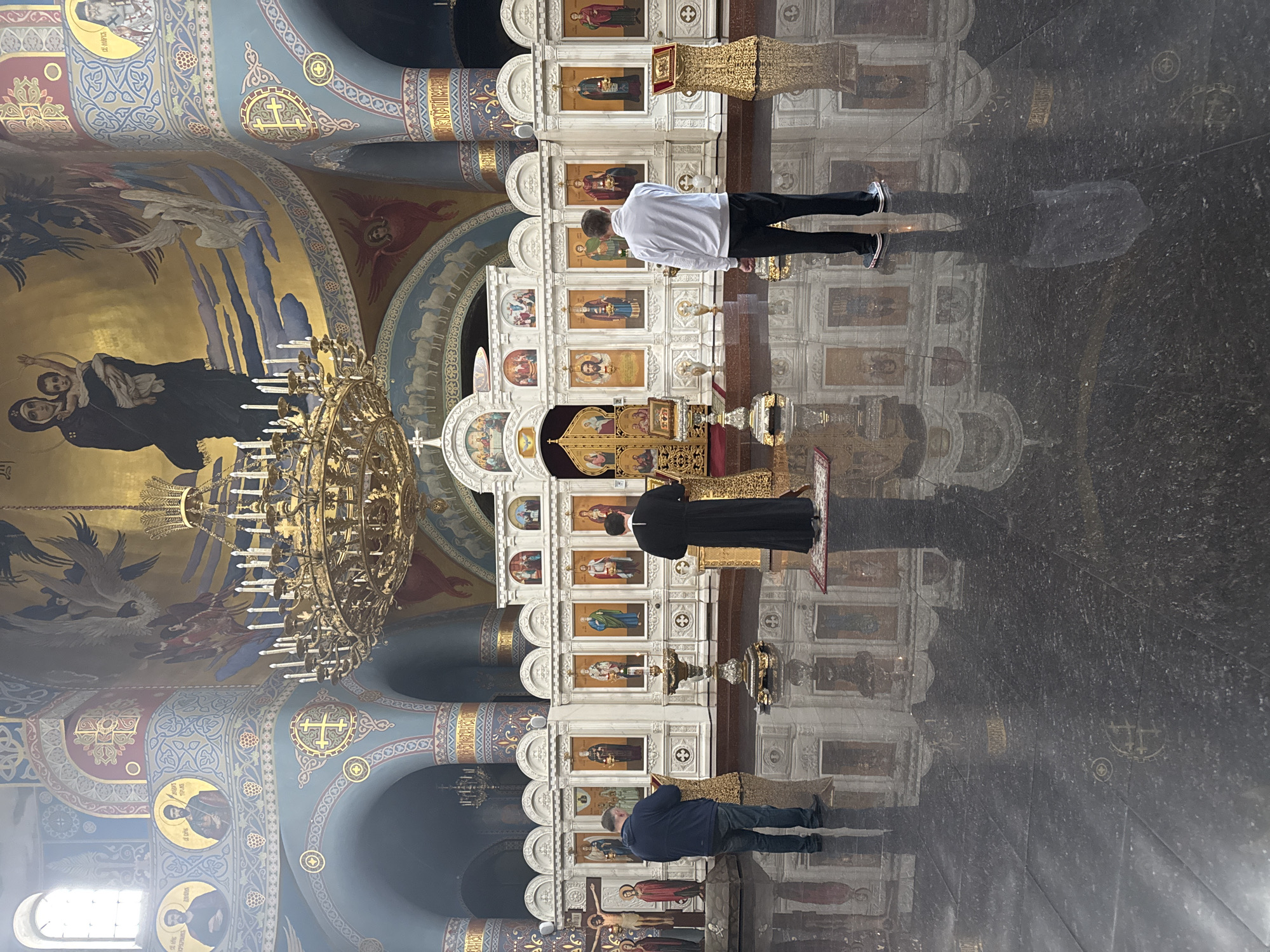 The image shows the interior of a church in Sochi, Russia. The walls and ceiling are adorned with religious paintings and gold accents. A large chandelier hangs from the ceiling. Three individuals are visible in the image. One person wearing a white shirt and black pants is standing in front of the altar, while a second person in a black robe is kneeling. Another individual wearing a blue jacket and jeans is standing near the front of the altar. The highly polished floor reflects the images of the people and the altar.