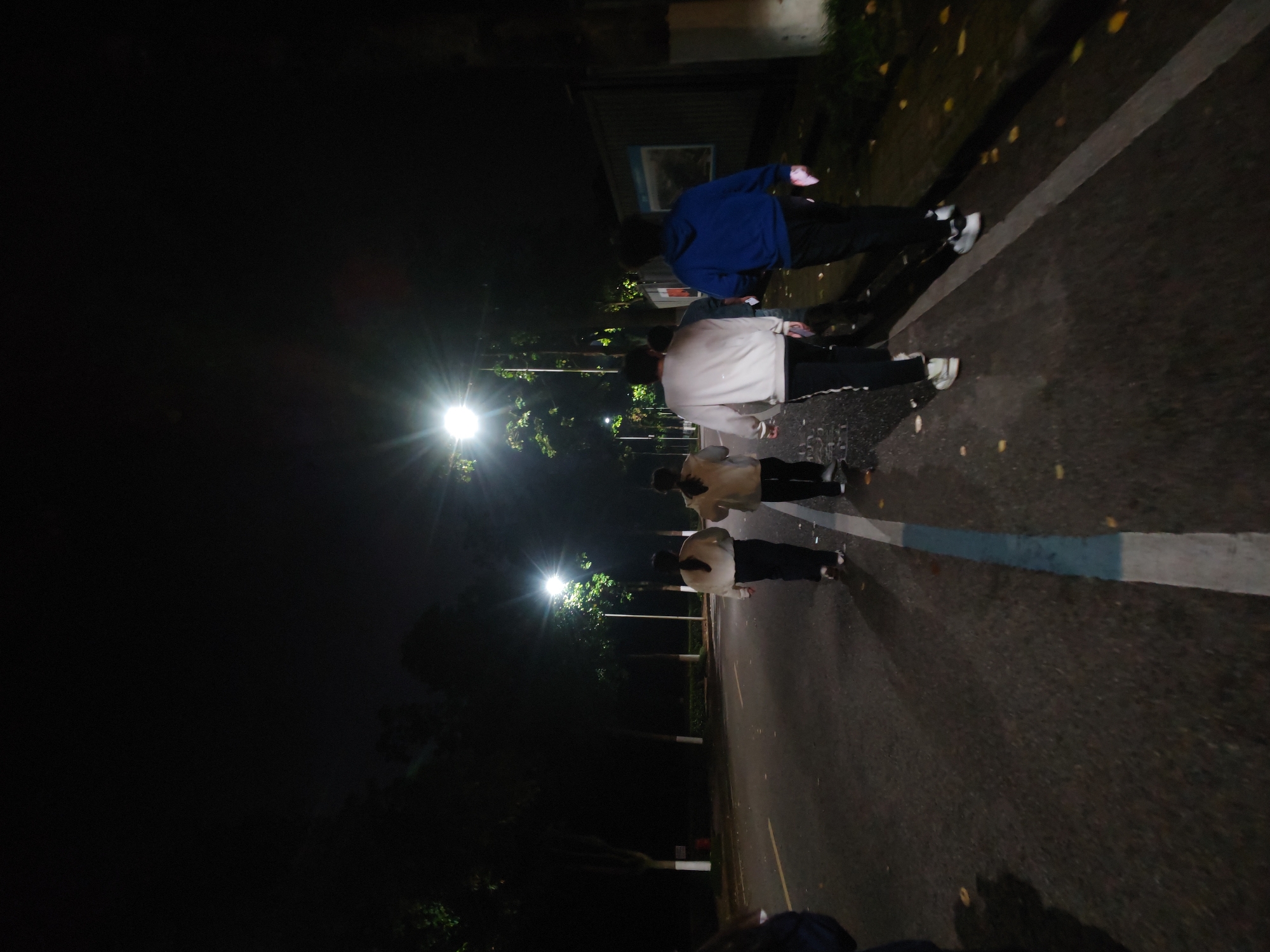 A dimly lit street scene is depicted at night in Chongqing High-tech Zone, China. The image is oriented sideways, with a group of four individuals walking along the pavement. The first person is wearing a blue hoodie with dark trousers. The second person in the sequence is wearing a light-colored jacket with dark pants. The third person has a light-colored jacket with dark pants. The last person has light-colored jacket with dark pants. The asphalt road is visible, marked with a white line. Streetlights cast a strong glow, creating a dark and contrasted environment. Trees line the edge of the road.