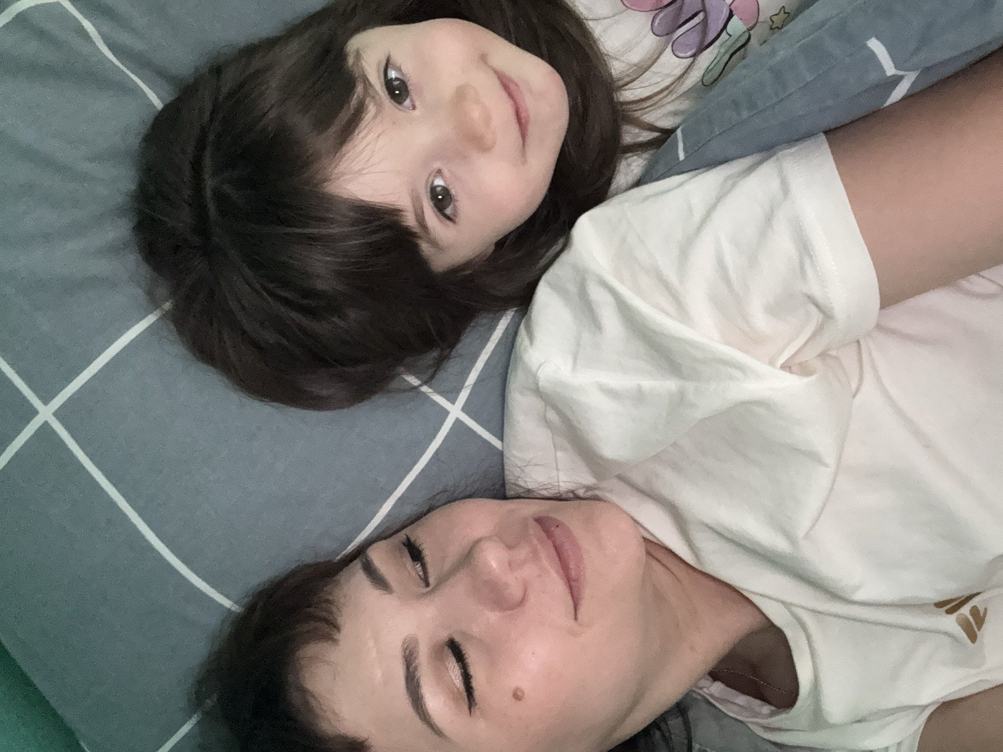 A high-angle medium shot shows a woman and a young child lying in bed, facing the camera. The woman, on the left, has her eyes closed and a slight smile on her face. She is wearing a light-colored t-shirt. The child, on the right, is looking directly at the camera with a happy expression and a smile. The child also has a t-shirt on. They are both lying on a bed with a gray and white checkered patterned duvet. The background is blurred, focusing attention on the two individuals.