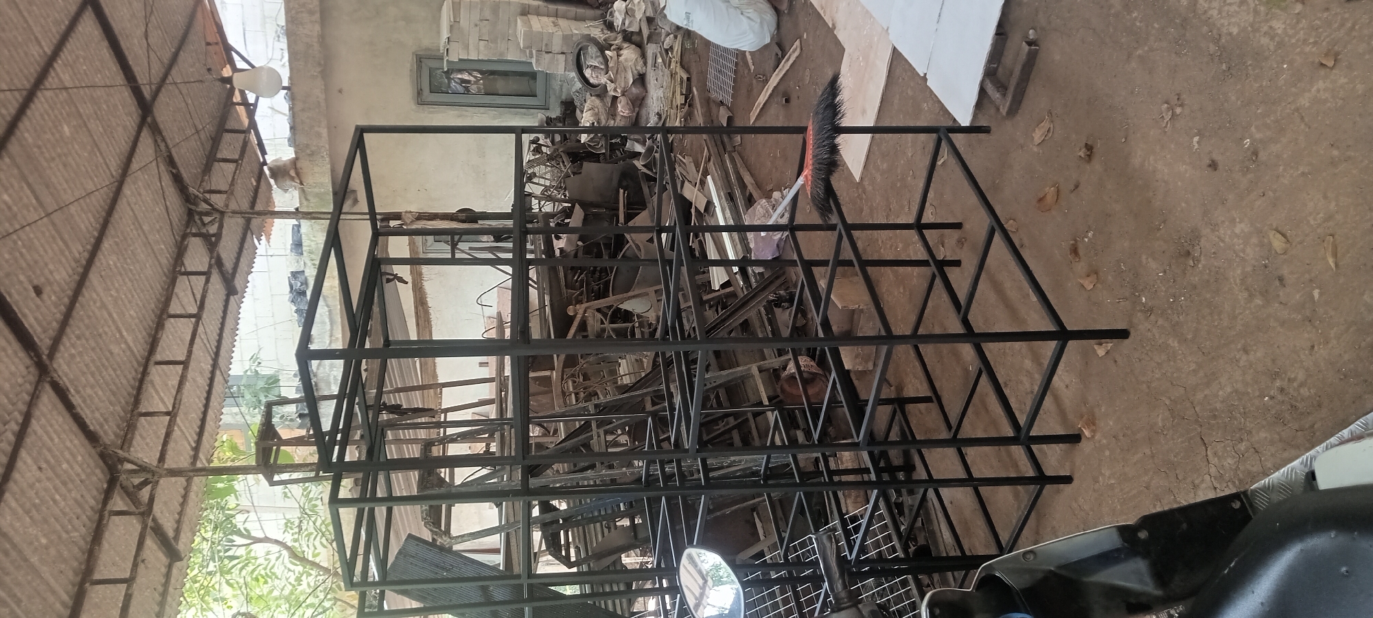 The image depicts a cluttered workshop area in Jogomulyo, Indonesia. The foreground is dominated by a large, black metal shelving unit, seemingly under construction. To the left, the front of a black motorcycle is visible, with its headlight illuminated. The ground is a dirt floor, scattered with dry leaves and some debris.
In the background, the workshop is filled with various metal objects, tools, and building materials. There are stacks of what appear to be metal bars, possibly scrap metal, and other disassembled furniture or equipment. A corrugated metal roof, supported by metal beams, covers the structure, suggesting an open-air or partially enclosed space. Light filters in from the sides, illuminating the scene. A broom with an orange handle and black bristles rests near the metal shelving. In the far right background, a person in a white garment is partially visible, along with a pile of sacks. A window and a stack of bricks are also present in the background wall. The overall impression is one of an active but disorganized workspace.