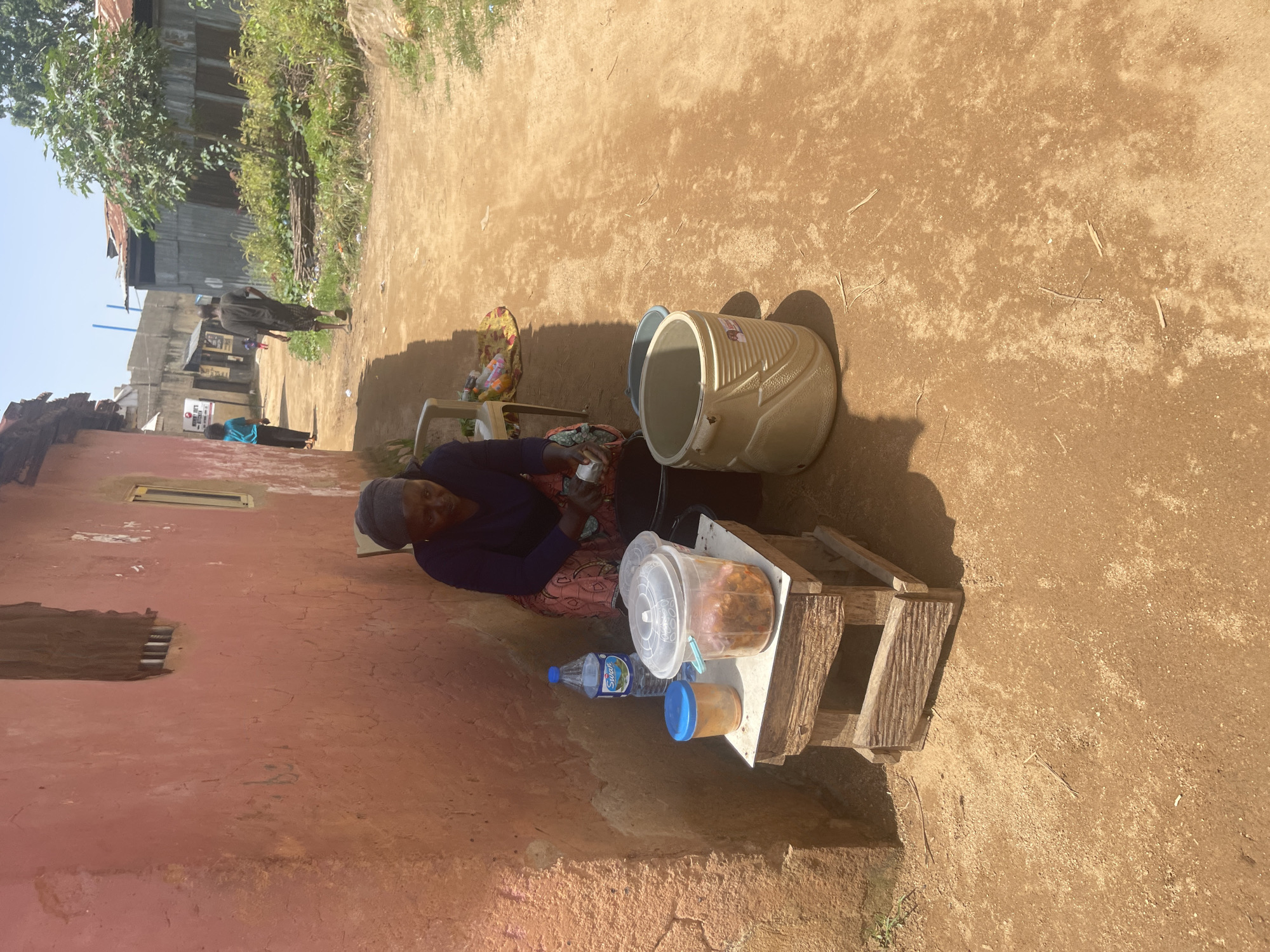 A woman in a dark headwrap and a purple top sits on a chair next to a pink plastered wall in what appears to be a market or residential area in Jos, Nigeria. In front of her, on a rustic wooden stand, are two containers of food and a bottle of water. To her right, on the ground, is a large beige insulated cooler and a smaller blue bucket. A dirt path runs alongside the wall, with other people visible in the background walking along the path. The area appears to be outdoors, with a bright, sunny sky. The wall of the building has a rough texture and two openings, one resembling a window with bars and the other a smaller rectangular opening. The ground is a reddish-brown dirt.