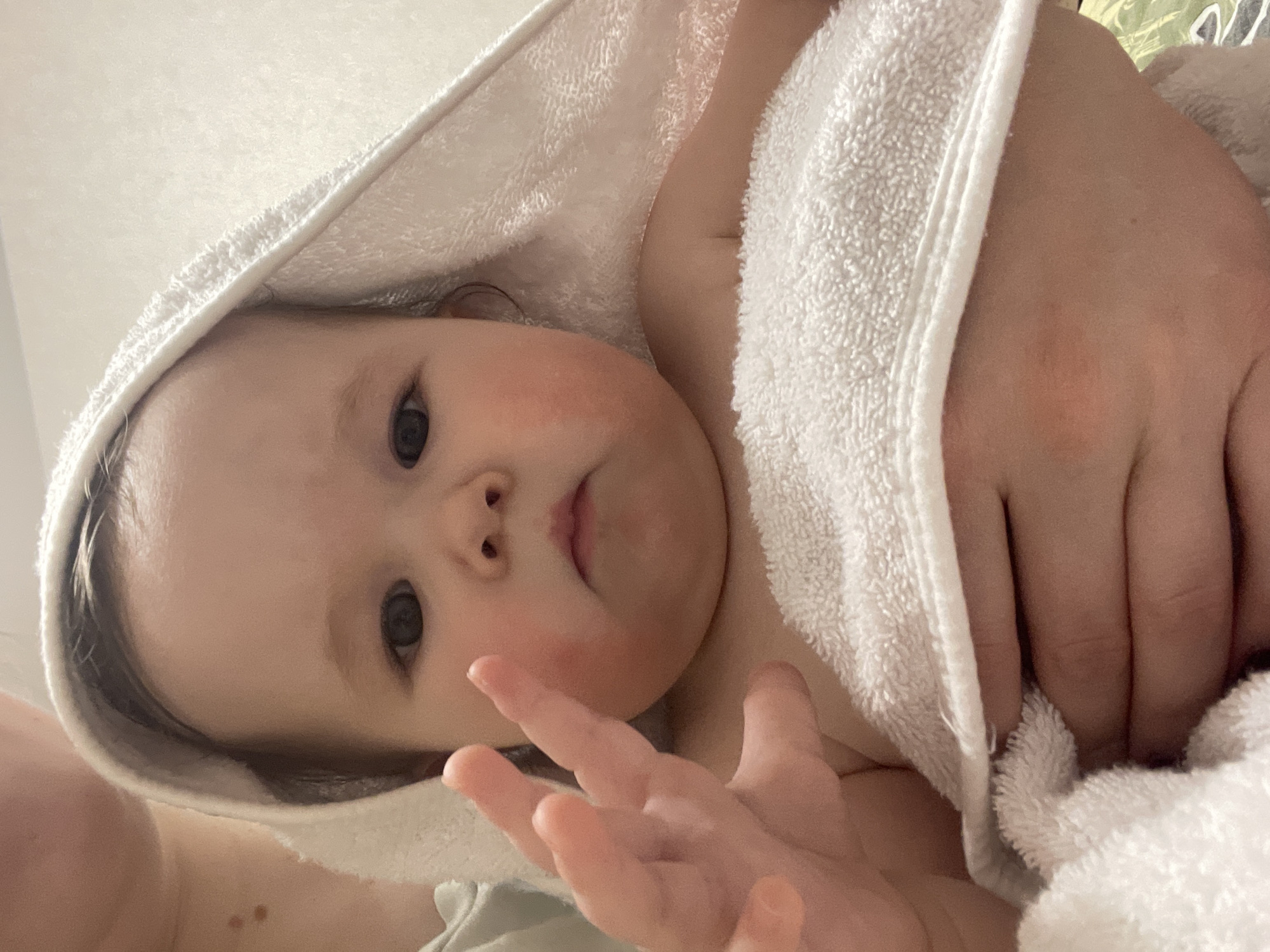 A close-up, upward-angled shot depicts a baby wrapped in a white towel, possibly after a bath. The baby's face, with slightly flushed cheeks, is the focal point, with their eyes looking directly at the camera. The towel is draped over the baby's head and around their body. Part of an adult's hand is visible, holding or securing the towel. The background is blurry and indistinct. There is no evidence that the picture was taken in Chaikovsky, Russia.