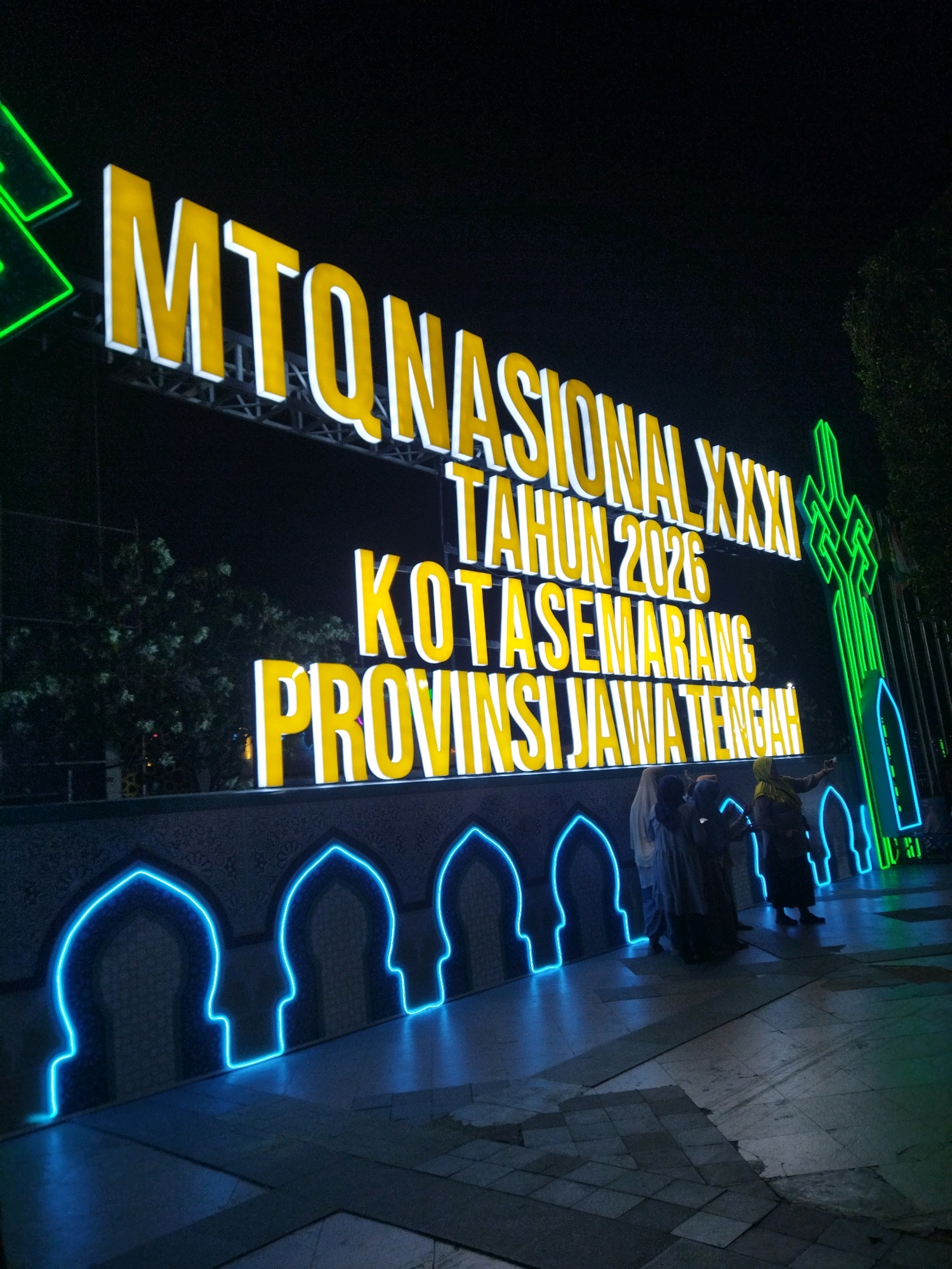 The image depicts a nighttime scene with a large, illuminated sign that reads "MTQ NASIONAL XXXI TAHUN 2026 KOTASEMARANG PROVINSI JAWA TENGAH." The sign is made of large, yellow, three-dimensional letters with white trim, set against a dark background. To the right of the sign is a tall, green, illuminated structure resembling a stylized minaret or abstract symbol. In front of the sign, there is a decorative border with arched shapes outlined in bright blue neon lights.
Several people, dressed in traditional Islamic clothing, are visible standing in front of the neon-lit border, some of whom appear to be taking photos. The ground in front of them is paved with stone tiles. The overall impression is that of an outdoor event or landmark in Semarang, Indonesia, celebrating the National Quran Recitation Festival in 2026.