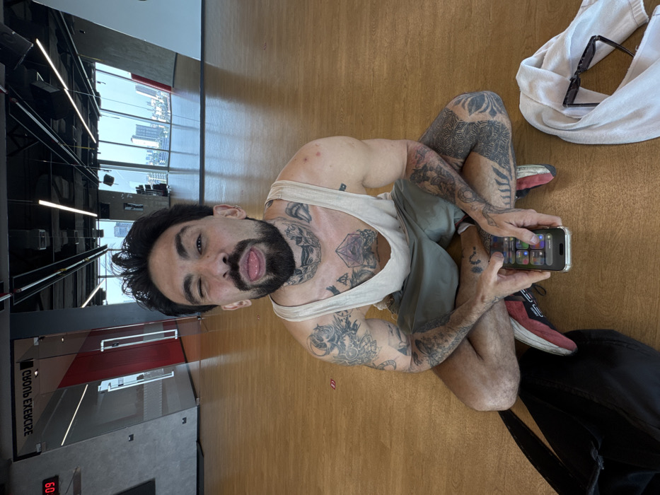 A male individual with dark hair, a beard, and extensive tattoos covering his arms, chest, and neck is seated cross-legged on a light brown wooden floor. He wears a beige tank top, green shorts, and sneakers with red accents. He is winking with his left eye and extending his tongue. He holds a silver-framed smartphone, displaying a user interface with various icons, in both hands. To his left lies a black backpack, and to his right, a white towel and dark sunglasses. The scene is an indoor exercise or fitness studio with large glass windows offering a panoramic view of a city skyline dominated by tall buildings under a bright sky, identifying the location as Bangkok, Thailand. The studio features grey wall panels, exposed ceiling structures with linear lighting, and reflective surfaces. A glass partition on the left has "GROUP EXERCISE" visible in reverse text and a digital clock displaying "09:".