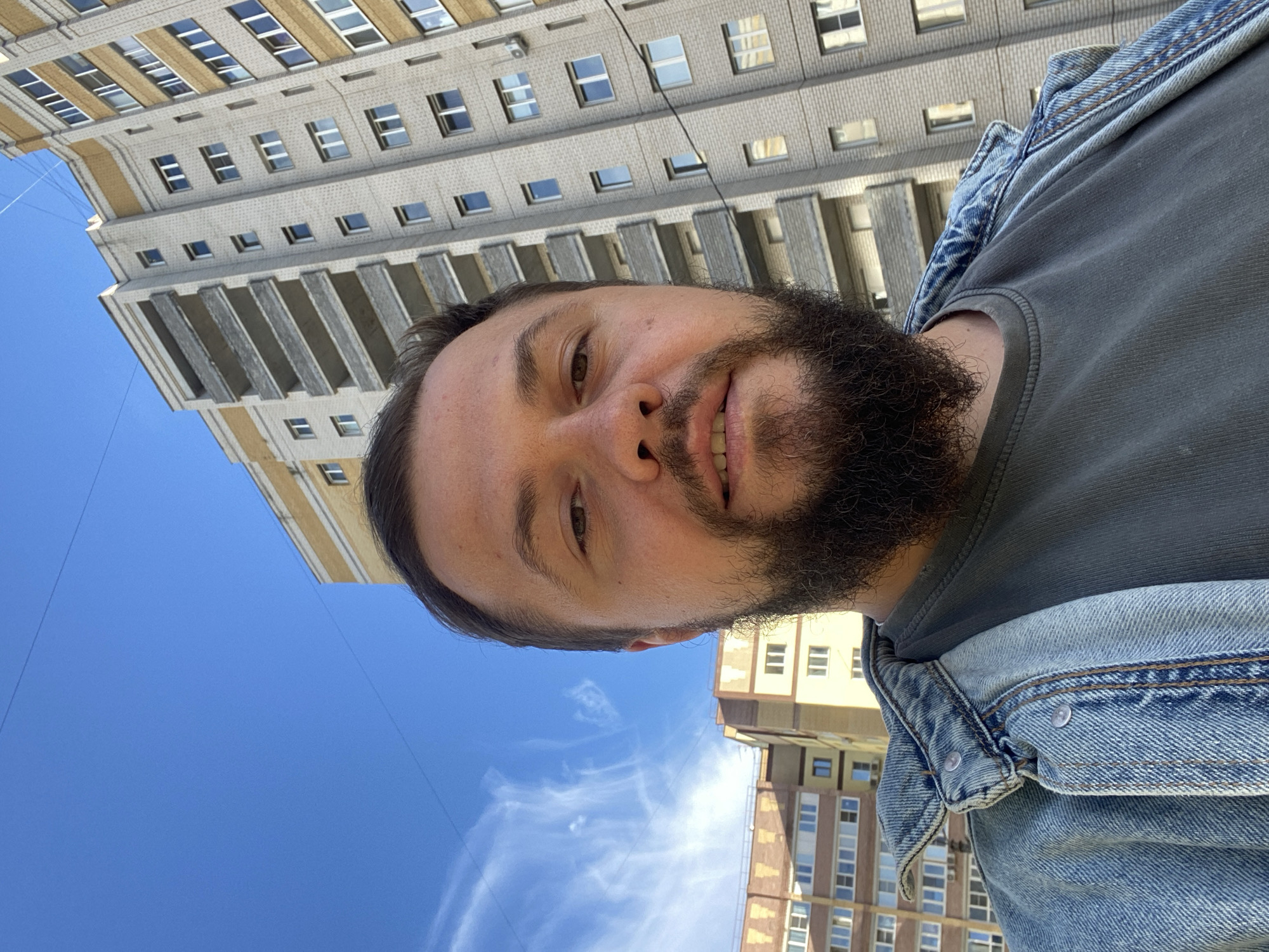 A medium shot selfie shows a man with a beard and brown hair standing outside a multi-story apartment building. He is wearing a grey t-shirt and a blue denim jacket. The building is made of light-colored brick with yellow accents and features multiple balconies. The sky is blue with some wispy clouds. The image appears to have been taken on a sunny day. The building in the background is typical of Soviet-era architecture found in many cities, including Vladimir, Russia.
