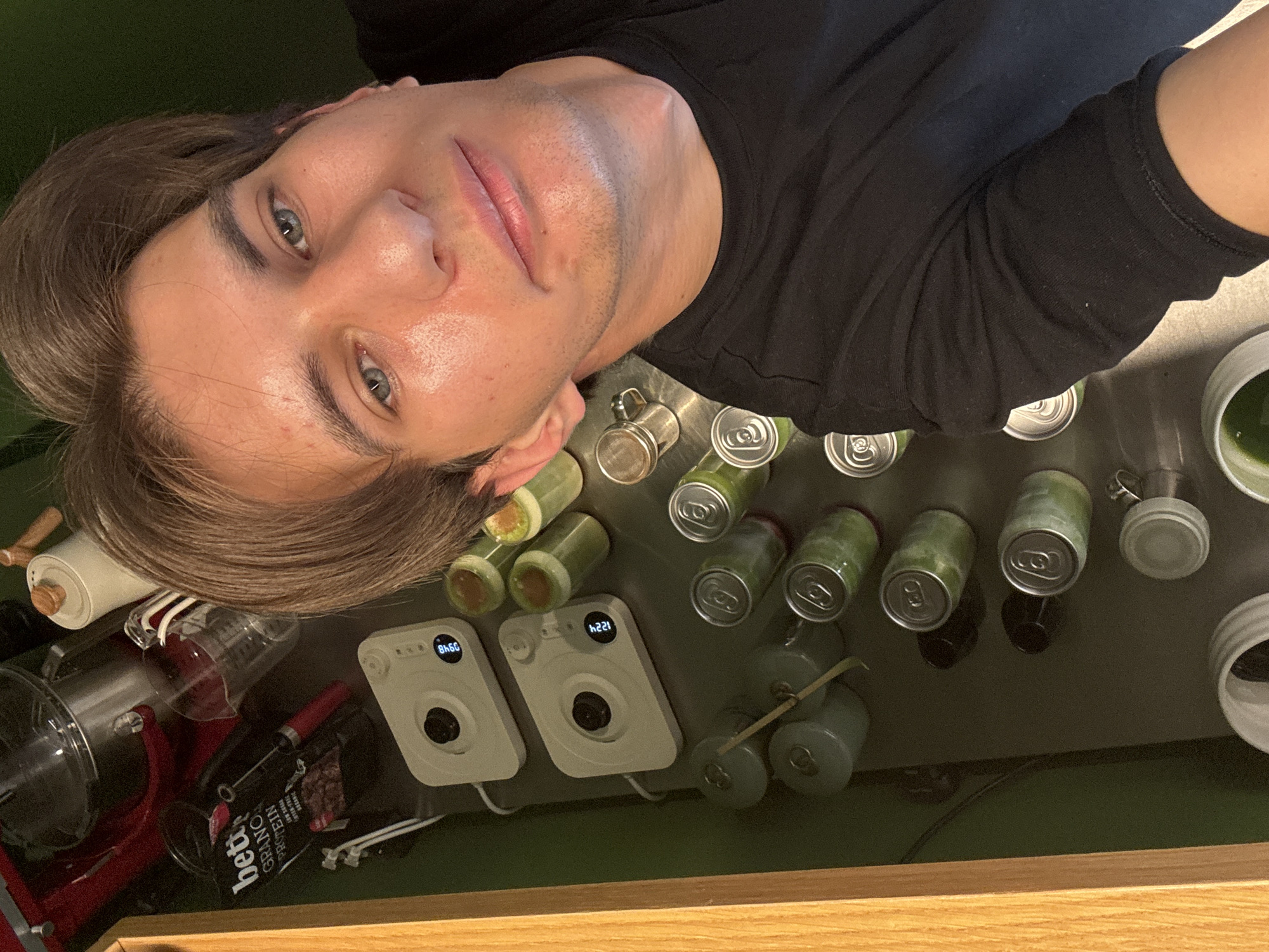 A man is pictured from an overhead perspective, likely a selfie. He has light brown hair, fair skin, and blue eyes, and is wearing a black long-sleeved shirt. He is positioned to the left of the frame.
To the right of the man is a countertop or table surface covered with various items:
- Several green cans and bottles arranged in rows.
- Two white electronic devices with circular features and digital displays, one reading "8460" and the other "1224".
- A red kitchen appliance (possibly a stand mixer) with accompanying attachments and a clear measuring cup.
- A container labeled "beta GRANOLA PROTEIN".
- Some small, green items with sticks protruding from them.
- A green surface below the countertop.
- A wooden surface near the bottom of the frame.
The location cannot be determined from the image. The city of Sofia, Bulgaria cannot be identified from this image.