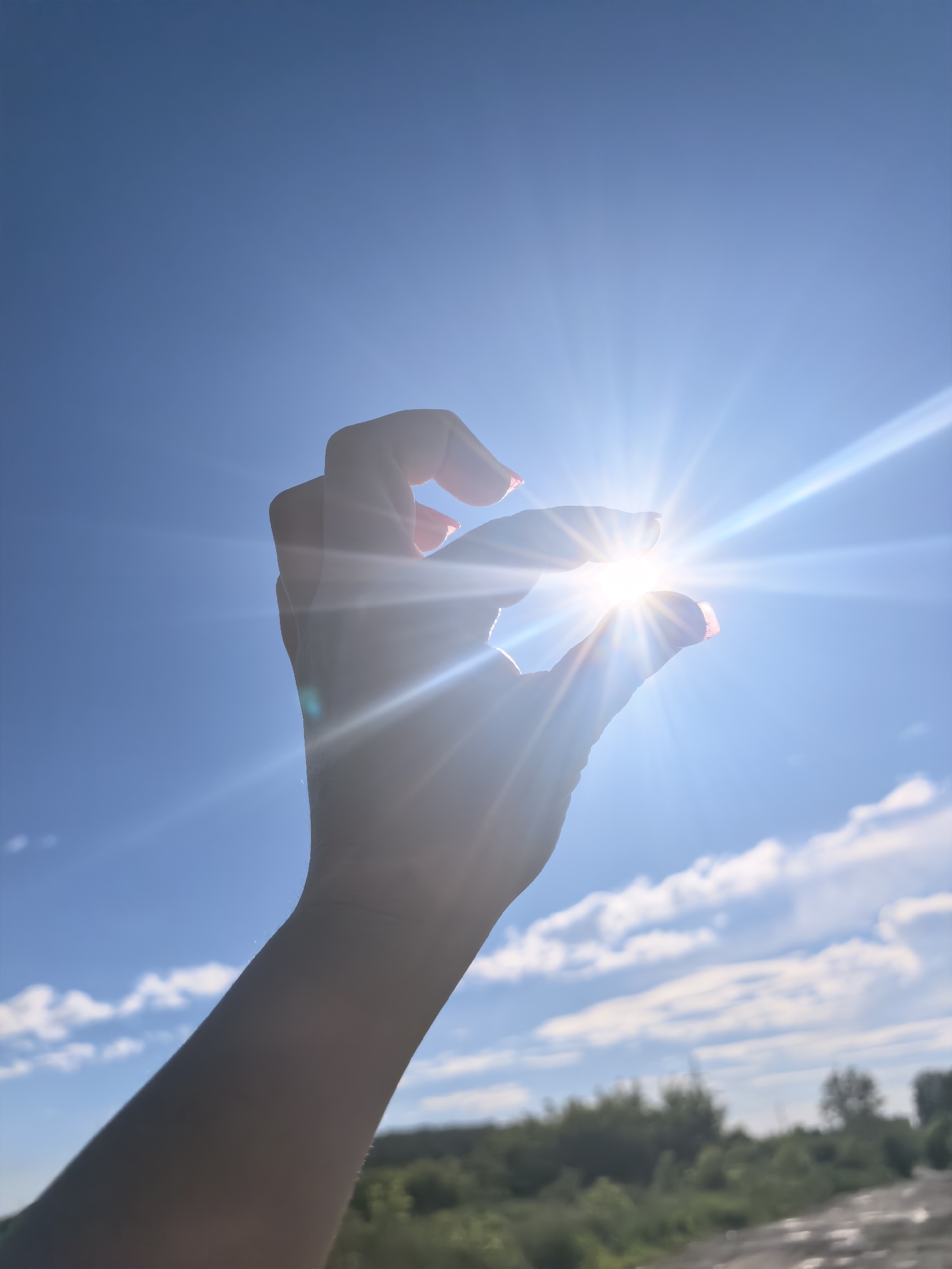 A hand is silhouetted against a bright blue sky, with the sun shining intensely through the gap between the thumb and forefinger. The sun's rays create a starburst effect, with bright streaks of light emanating outwards. The hand appears to be reaching up, as if trying to grasp the sun. In the background, there is a blurred landscape of green foliage and a few wispy clouds. The image is taken from a low angle, looking upwards towards the sky.