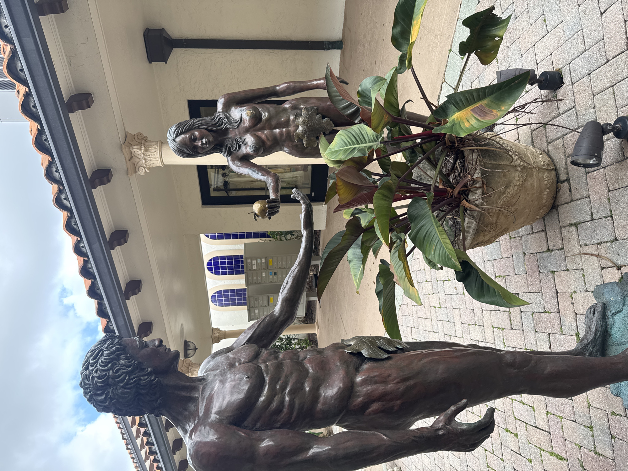 This image depicts a bronze sculpture of Adam and Eve situated in an outdoor paved courtyard, characteristic of the Mediterranean Revival architecture prevalent in Boca Raton, United States of America.
The male figure, Adam, stands on the left, facing right towards Eve. He is rendered with a muscular physique, short curly hair, and a somber expression. His right arm is extended forward, with his palm open, positioned as if to receive an object. A bronze leaf modesty covers his groin.
The female figure, Eve, stands on the right, facing left towards Adam. She is depicted with a slender form, long wavy hair, and a subtle smile. She holds a small, golden apple in her outstretched left hand, offering it towards Adam. A bronze leaf modesty covers her groin.
The ground beneath the statues is paved with light-colored, rectangular paver bricks, displaying minimal moss growth in some crevices. Between the two figures, a large, rustic-textured concrete planter holds a plant with broad, glossy leaves, primarily deep green with reddish-maroon undersides and stems. Two black, adjustable landscape spotlights are positioned on the pavers near the planter.
The background features a building with light-colored stucco walls and a terracotta barrel-tiled roof. A dark rain gutter system runs beneath the roofline, supported by decorative dark corbels. A covered walkway, or portico, extends along the building, supported by light-colored columns with ornate Corinthian capitals. In the distance behind Adam, several arched windows with blue glass panes are visible, with a bank of mailboxes or parcel lockers directly below them. Behind Eve, a black-framed glass doorway reveals clothing hanging inside, indicating a retail or boutique space. The sky above is partially cloudy.