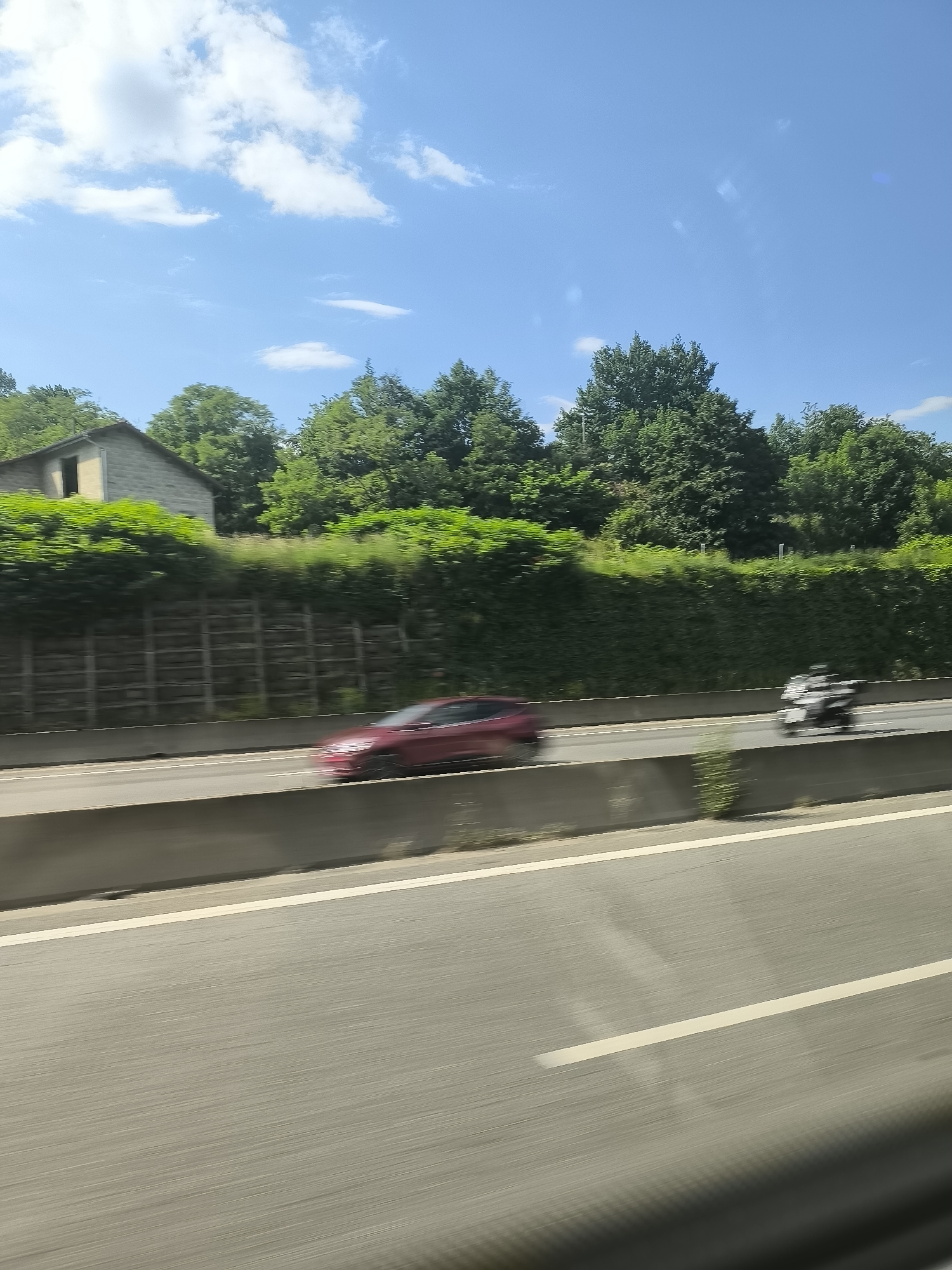 The image, taken from a moving vehicle, depicts a highway scene near Saint-Chamond, France. The sky is blue with scattered clouds. Along the road are trees and a building. A blurred red car and a motorcycle are moving on the highway. The focus is on the road and the passing vehicles.