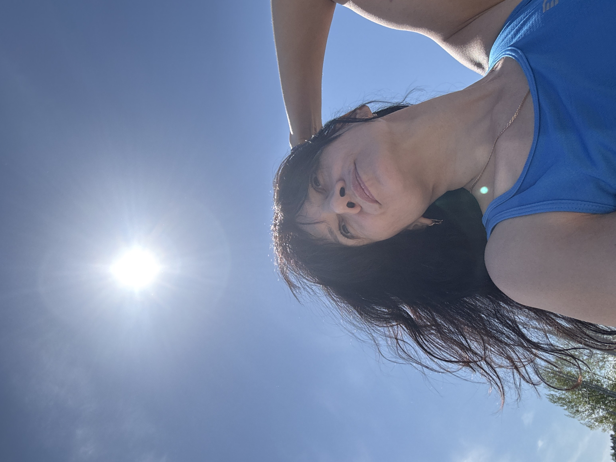The image is an upward-angled selfie of a woman with dark hair wearing a blue tank top and a gold necklace. Her arm is raised above her head. The background is a bright blue sky with the sun visible in the upper left corner. The location is possibly the city of Chaikovsky, Russia. The woman is smiling slightly. A few green trees are visible in the lower right corner.
