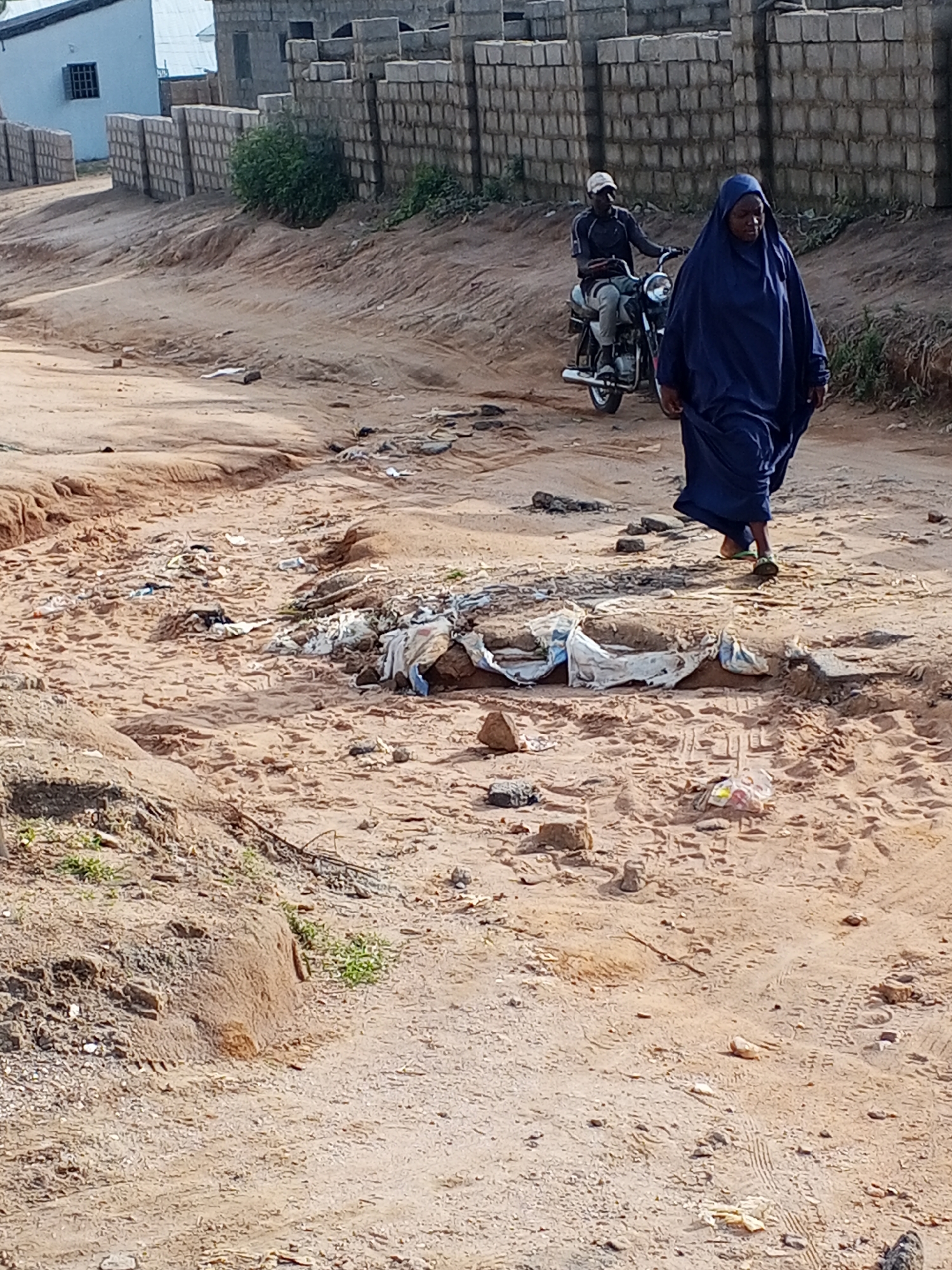 The image depicts a dirt road in what appears to be a residential area of Adonkolo, Nigeria. The road is heavily rutted and strewn with trash. A woman in a blue robe is walking along the road. In the background, a man on a motorcycle is riding in the same direction. To the side of the road is a partially constructed brick wall.