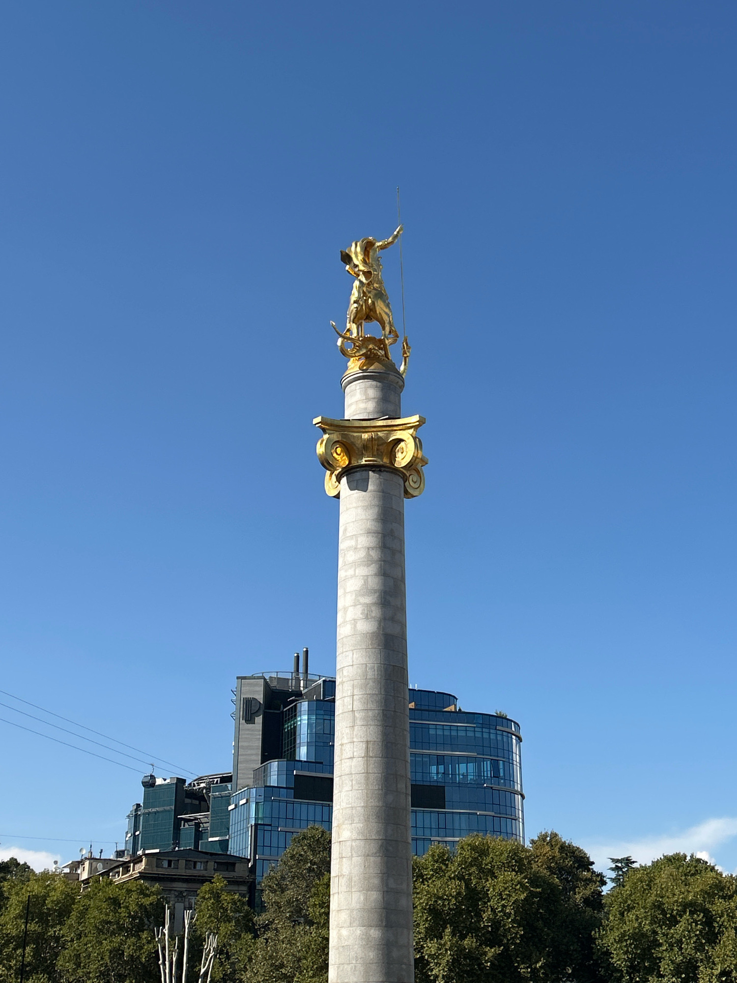 This is a medium-shot, eye-level, outdoor, vertical photograph taken on a clear day in Tbilisi, Georgia. Dominating the frame is the Narikala Fortress, a prominent landmark in the city. A golden statue of St. George is perched atop a tall, gray stone column, a symbol of Georgia's patron saint. The statue depicts St. George on horseback, spearing a dragon. The column is adorned with ornate, golden Corinthian capitals. In the background, modern glass-fronted buildings and lush green trees can be seen, interspersed with a cable car gliding across the skyline. The sky is a vibrant, cloudless blue.