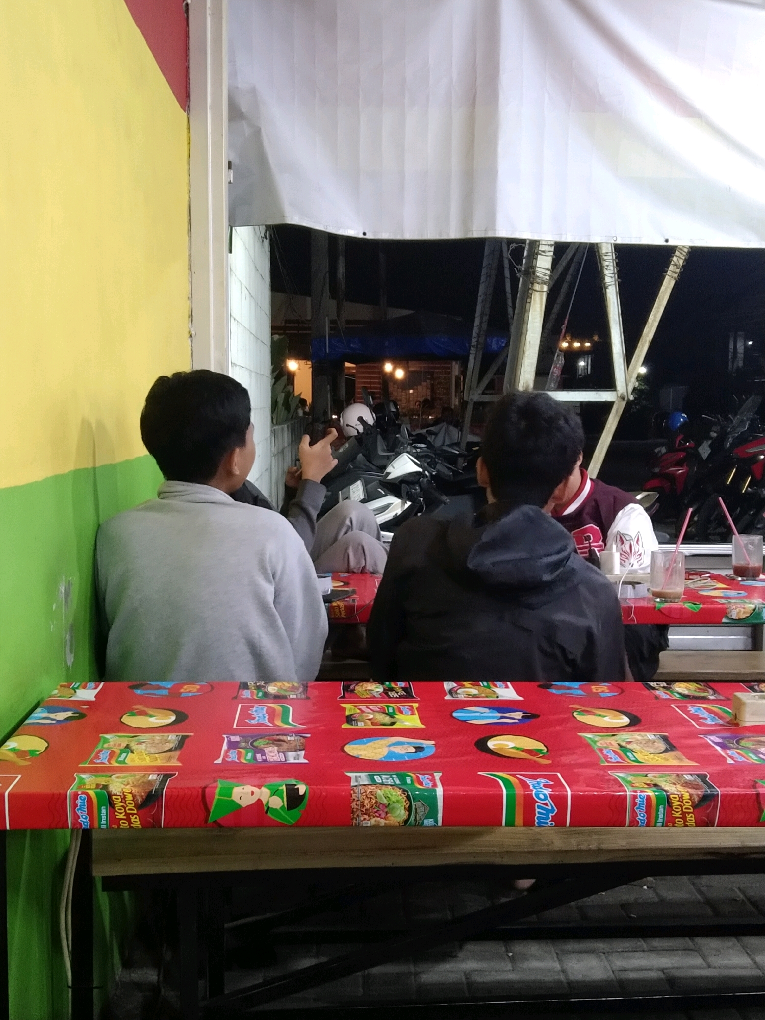 Two young males sit at a red table covered with a tablecloth printed with images of instant noodle packaging and cartoon figures. The table is set up outdoors, possibly at a street-side eatery in Malang, Indonesia. The individual on the left, facing away from the camera, wears a light gray hooded sweatshirt and sits near a yellow and green wall. The individual on the right, also facing away, is wearing a dark jacket and is looking towards a person in a maroon and white jacket who is partially visible. A motorcycle is parked behind them, and the scene appears to be at night, indicated by the artificial lighting and dark background.