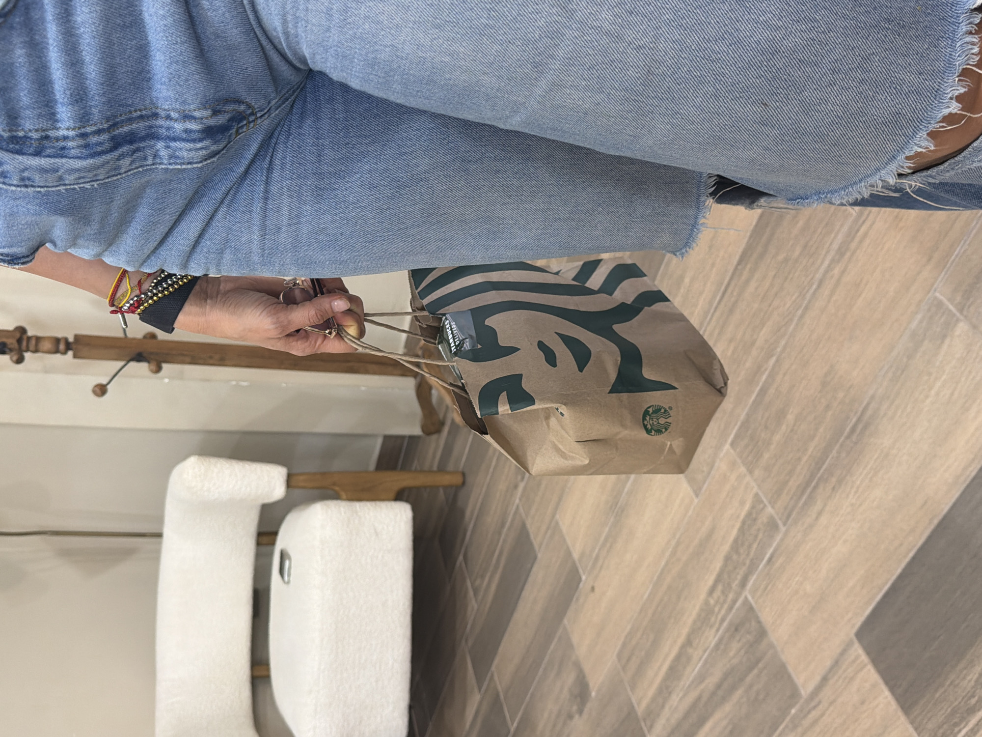 The image shows a person wearing light blue denim jeans, holding a brown paper Starbucks bag with green logos and handles. The person is wearing multiple bracelets on their left wrist. The background includes a light-colored tile floor in a herringbone pattern, a white upholstered bench or ottoman, and a wooden clothing rack. The location is likely indoors, but more context is needed to confirm the city of Villahermosa, Mexico.