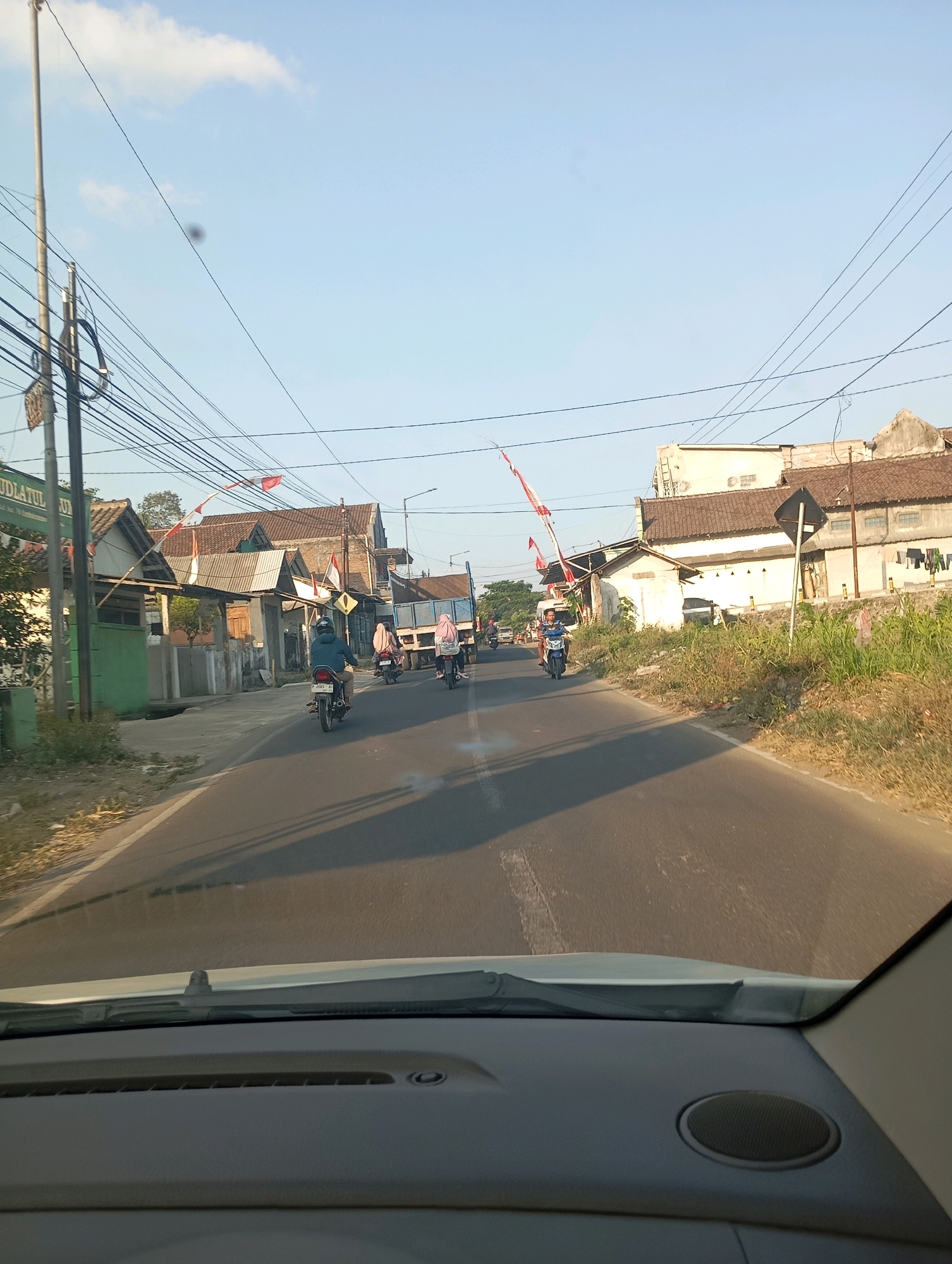 The image depicts a street scene in Slateng, Indonesia, likely during daytime given the clear sky and sunlight. The perspective is from inside a vehicle, showing the dashboard in the foreground. The road is paved and appears to be a two-lane street with some traffic.
Several individuals are present, primarily on motorcycles. In the middle of the frame, a person is riding a motorcycle in the left lane, facing away from the camera. Further ahead, two more motorcycles are visible, one with a rider and a passenger wearing headscarves, and another with a single rider. On the right side of the road, a light blue truck is driving in the same direction as the motorcycles.
The environment includes buildings lining the street, with a mix of residential or small commercial structures. Some of these buildings have tiled roofs. There are also numerous power lines and utility poles running along the street, indicating an urban or semi-urban setting. Vegetation, including grass and some trees, is visible on the sides of the road. Indonesian flags are also present, some attached to poles along the street and one on a pole in the distance. The overall atmosphere appears to be a typical day in a populated area with active street life.