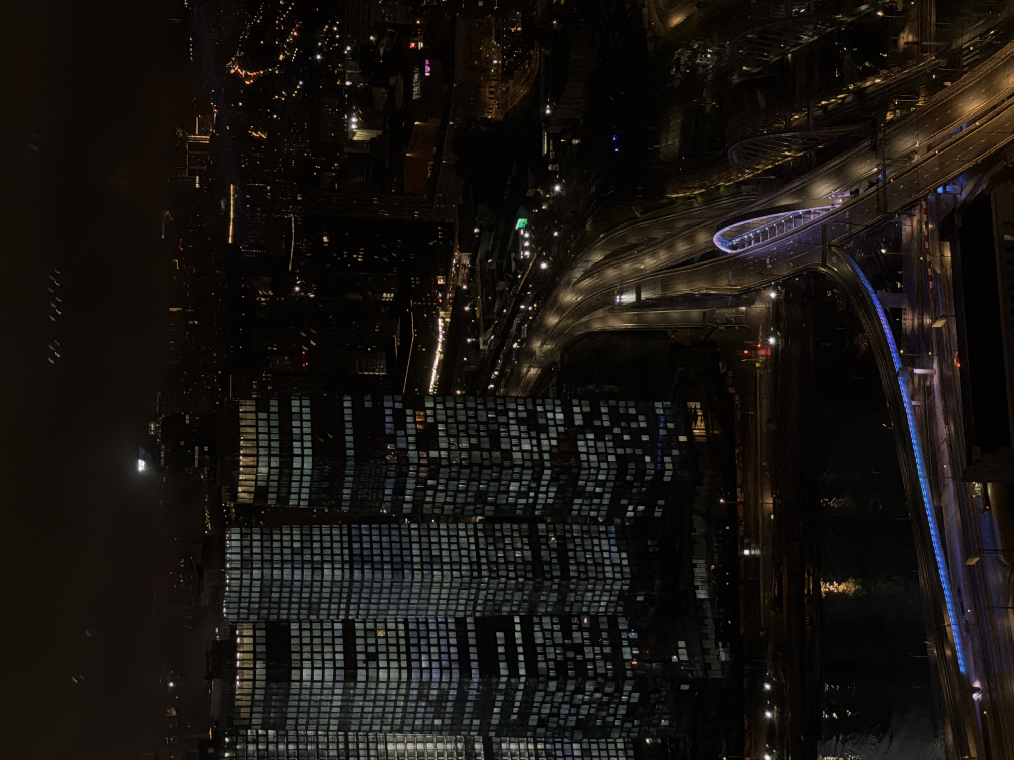 The aerial, high-angle, night-time photograph depicts a section of the Moscow Oblast, near Khimki, Russia, characterized by a dense urban landscape illuminated by artificial lights. Dominating the foreground are three prominent, modern skyscrapers with grid-like patterns of lit windows, suggesting occupancy. Adjacent to these structures, a multi-lane highway curves through the scene, with vehicle headlights and taillights creating streaks of light on the wet-looking asphalt. Further into the background, the urban sprawl continues with a multitude of smaller buildings and distant city lights that fade into the dark sky. A prominent bridge with distinctive blue and purple lighting spans across a body of water in the lower portion of the image, reflecting some of the surrounding lights. The overall atmosphere is one of a vibrant, active city at night, with the infrastructure of transportation and residential/commercial buildings forming a complex network. No individuals are clearly discernible in the image, only the presence of infrastructure and artificial illumination.