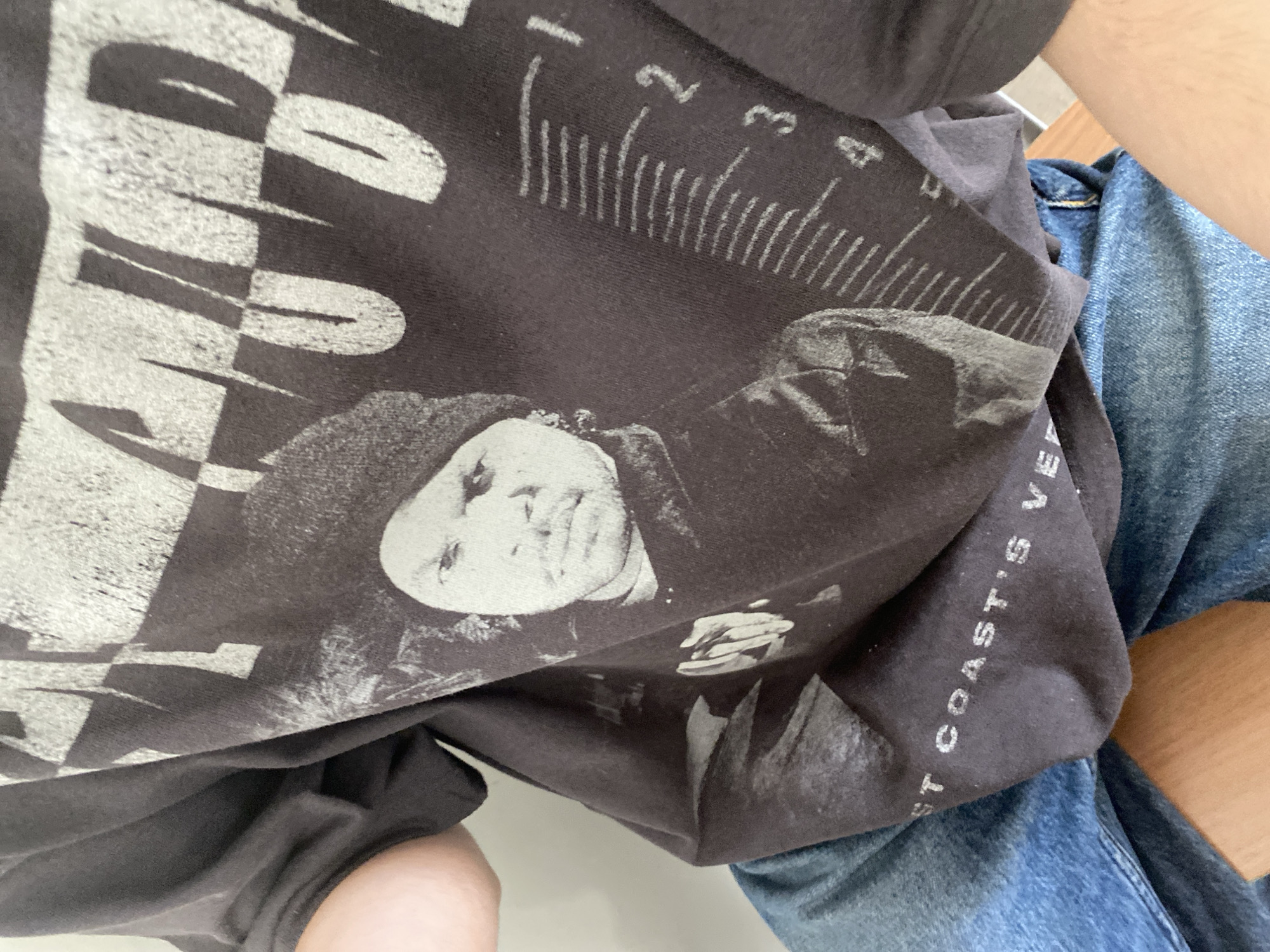 The image provides a close-up, high-angle view of a person seated, wearing a dark gray graphic t-shirt and blue denim jeans. Parts of the person's arms and legs are visible. A light-colored wooden surface is also visible on the right side of the frame.
The t-shirt features a prominent black and white graphic. It includes a portrait of a person, likely male, with a serious expression, looking slightly upward and to the right. The person in the graphic appears to be wearing some form of headwear, possibly a hat or beanie. To the left of the portrait, large, distressed white text is partially visible, spelling out "AND D..." (the rest is cut off), set against a checkerboard or striped pattern. Above the portrait, there is a ruler-like graphic with white markings, including numbers "1", "2", "3", and "4", and numerous vertical tick marks. Along the bottom right curve of the t-shirt, smaller white text reads "...ST COAST'S VE...", suggesting a phrase like "West Coast's Very Best" or similar, related to a geographical region. The t-shirt fabric is creased and shows signs of wear, indicating a casual style. The scene depicts a relaxed, informal setting, with no specific activity occurring other than the person being seated. The location context of Yerevan, Armenia, is not visually discernible from the image content.