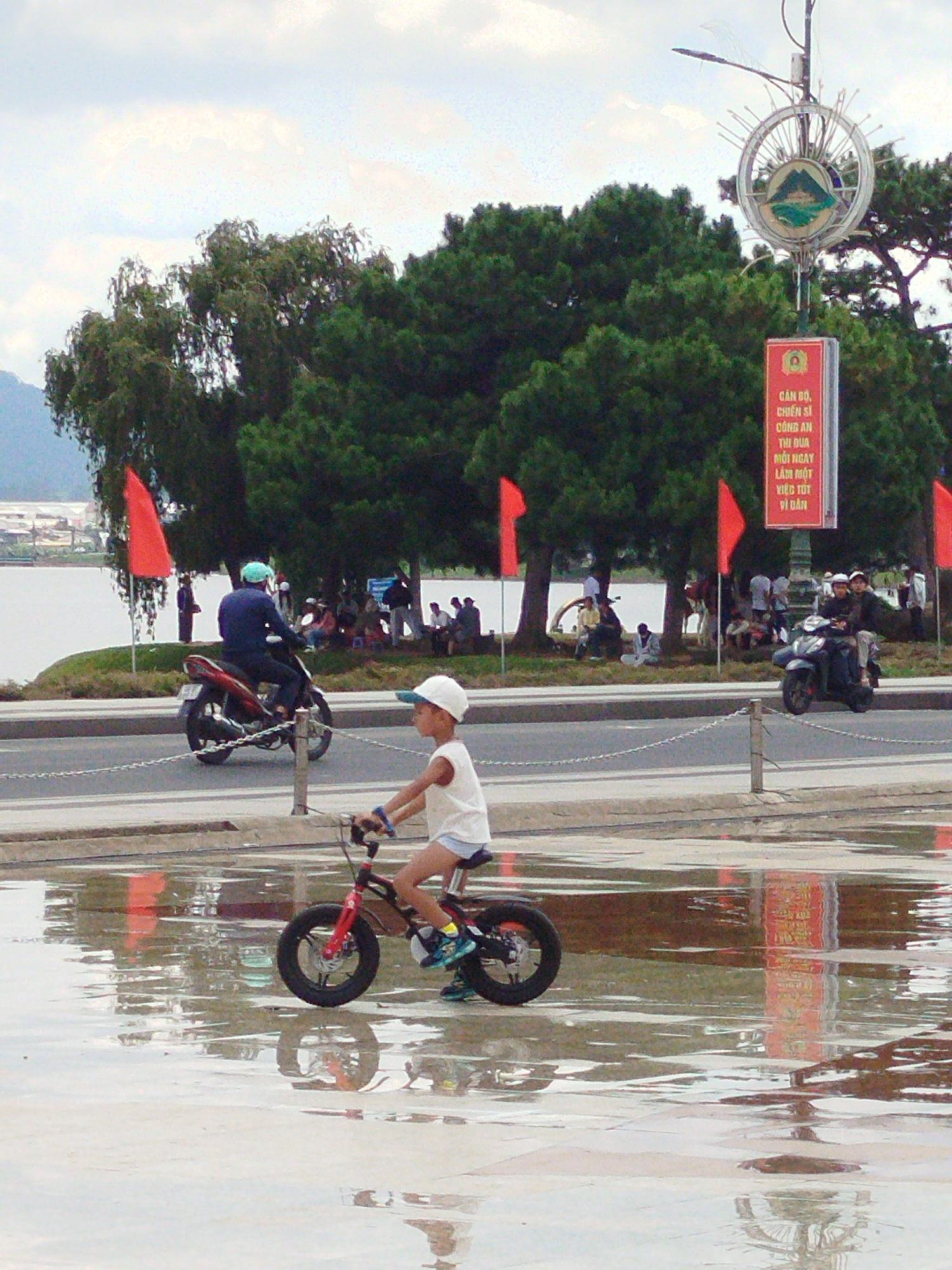 In the foreground, a young child wearing a white cap and a light-colored shirt rides a small bicycle through a large puddle reflecting the surrounding environment. The bicycle has a red front fork and black wheels. Further in the background, a person wearing a blue jacket and a light blue helmet rides a red and black scooter. The scooter's rider is looking towards the left. Behind the scooter, several people are gathered under trees. A tall pole with a circular emblem and a red banner with Vietnamese text stands near the gathered people. Red flags are visible on poles to the left and right of the scene. In the distance, a body of water, likely Xuan Huong Lake, is visible, with buildings and hills on the far side. The sky is cloudy. This scene is located on a road adjacent to Xuan Huong Lake in Da Lat, Vietnam, identifiable by the presence of the lake, the surrounding hills, and the specific architectural and cultural elements, such as the banner.