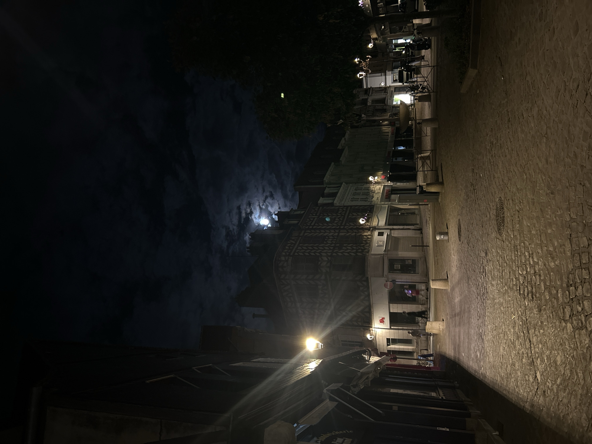 The image depicts a nocturnal view of a cobblestone street in Auxerre, France, likely within the historic city center. The full moon is visible in the dark sky, partially obscured by clouds, casting a soft illumination on the scene. The street is lined with traditional French buildings, some featuring timber-framed facades. Streetlights are present, with one casting a strong, directional beam down the pathway.
In the foreground, the cobblestone pavement is wet, reflecting the light. There are a few people walking on the street; one individual is riding a bicycle, and another appears to be walking. Several concrete bollards are placed along the side of the street. In the mid-ground, there are shopfronts, some with awnings extended. A water fountain is visible in the distance. The overall atmosphere is quiet and serene, characteristic of a city at night.