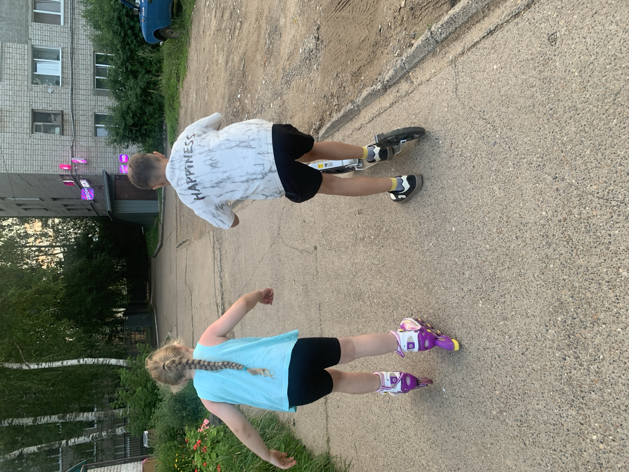 Two children are engaged in recreational activities on a paved walkway in Chaikovsky, Russia. On the left, a young girl with blonde hair in a braid is wearing a light blue tank top, black shorts, and purple roller skates. She is facing away from the camera and appears to be in motion. On the right, a boy, also facing away, is riding a scooter. He is wearing a white t-shirt with "HAPPINESS" printed on the back, black shorts, and black and white sneakers with yellow socks.
The environment suggests a residential area, with a multi-story brick building visible in the background. The building has windows and balconies, and what appear to be business signs with neon lighting. Trees and green foliage surround the building and the walkway. A blue vehicle is parked to the right of the building. The walkway itself is made of concrete with some debris and tire marks visible on the adjacent dirt or gravel area. The lighting suggests it is either early morning or late afternoon.