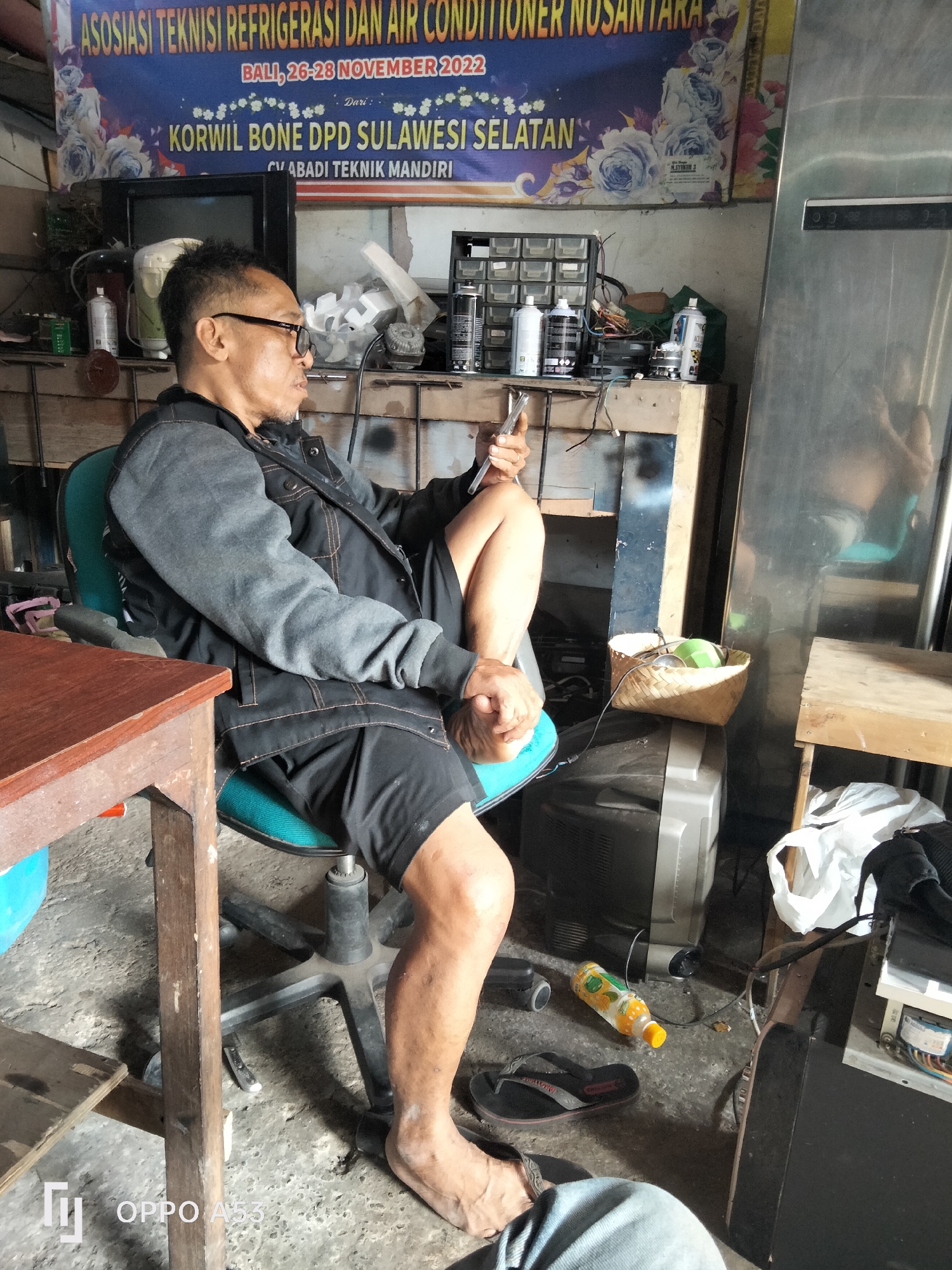 The image captures an indoor scene, likely a workshop or storage area, in Denpasar, Indonesia. A man, wearing glasses, a dark jacket, and black shorts, sits in a turquoise office chair, holding a small object in his right hand and looking to his right. His left arm is resting on the armrest of the chair. The environment is cluttered with various items, including a vintage television set, a large stainless steel refrigerator, storage containers filled with small parts, a basket containing a green object, various tools, and electrical wiring. A blue chair is partially visible to the left. A wooden table with a reddish-brown top is positioned in front of the man. The floor is concrete and appears dusty. In the background, a banner with text and floral imagery is hanging on the wall, indicating an event related to "ASOSIASI TEKNISI REFRIGERASI DAN AIR CONDITIONER NUSANTARA" held in Bali from November 26-28, 2022, with "KORWIL BONE DPD SULAWESI SELATAN" and "CV ABADI TEKNIK MANDIRI" also mentioned. A bottle of orange-colored liquid and a pair of black flip-flops are on the floor. The overall impression is of a utilitarian space, possibly a repair shop, where the man is taking a break.
