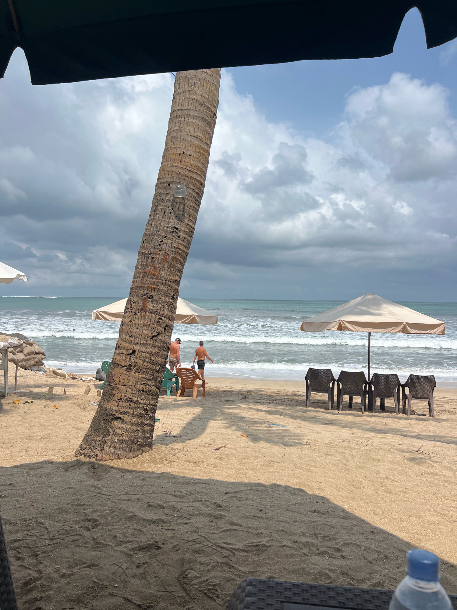 A beach scene in Kuta, Indonesia, with a large, textured palm tree trunk dominating the foreground on the left. The sandy beach extends to the right and meets the ocean, which has moderate waves. Two men are visible on the beach, one walking away from the camera, and the other facing the ocean. Several beach chairs and umbrellas are set up on the sand. The sky is cloudy with patches of blue. The overall atmosphere is relaxed and typical of a tropical beach destination.