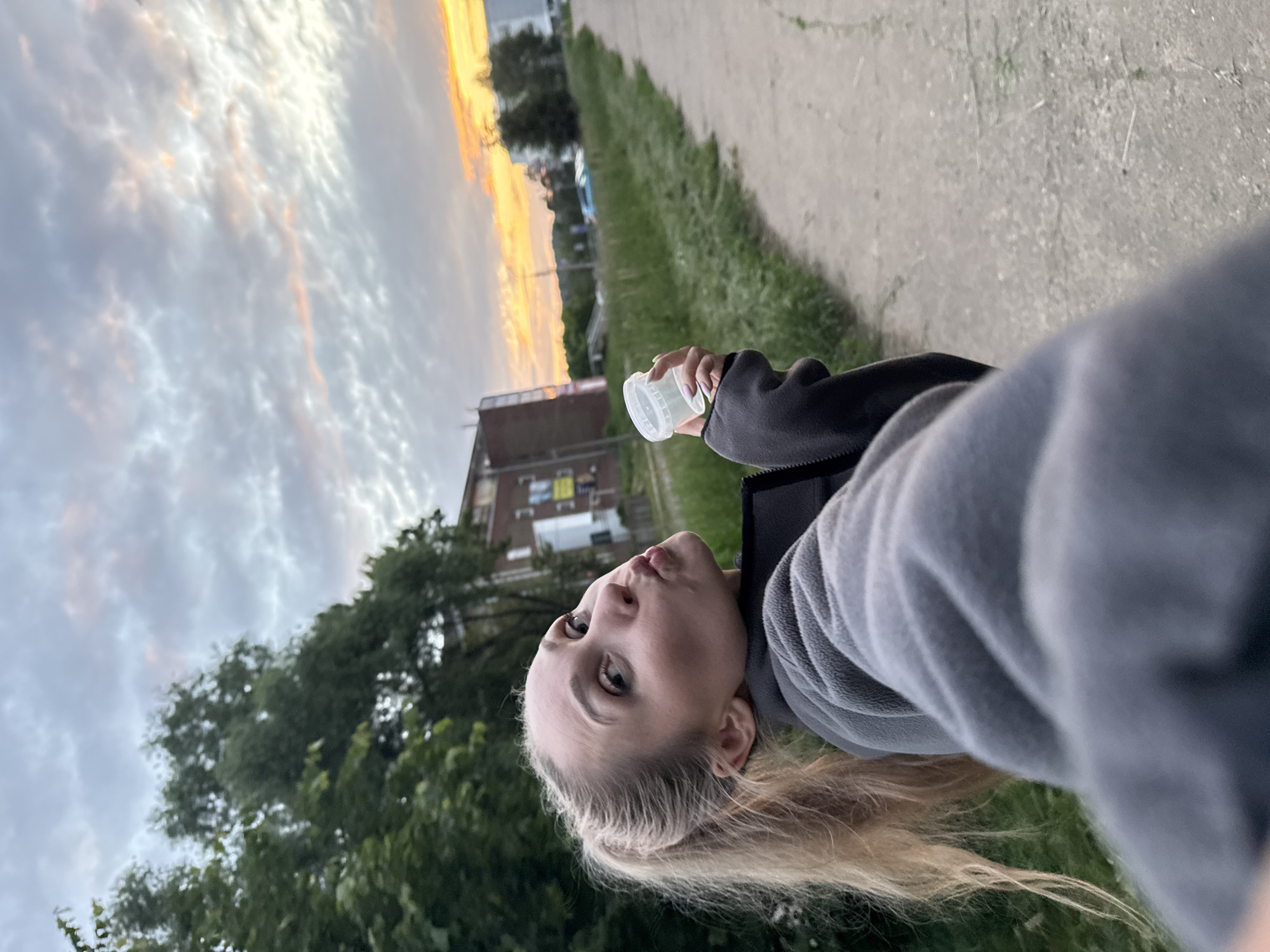 A young woman with blonde hair pulled into a ponytail, wearing a grey fleece jacket, takes a selfie outdoors in Chaikovsky, Russia. She is pouting her lips and holding a small white plastic container in her right hand. The background shows a paved path, a grassy area, trees, and a multi-story brick building. The sky is cloudy with hints of orange and yellow from a sunset. The scene appears to be a casual outdoor moment, possibly during an evening walk.