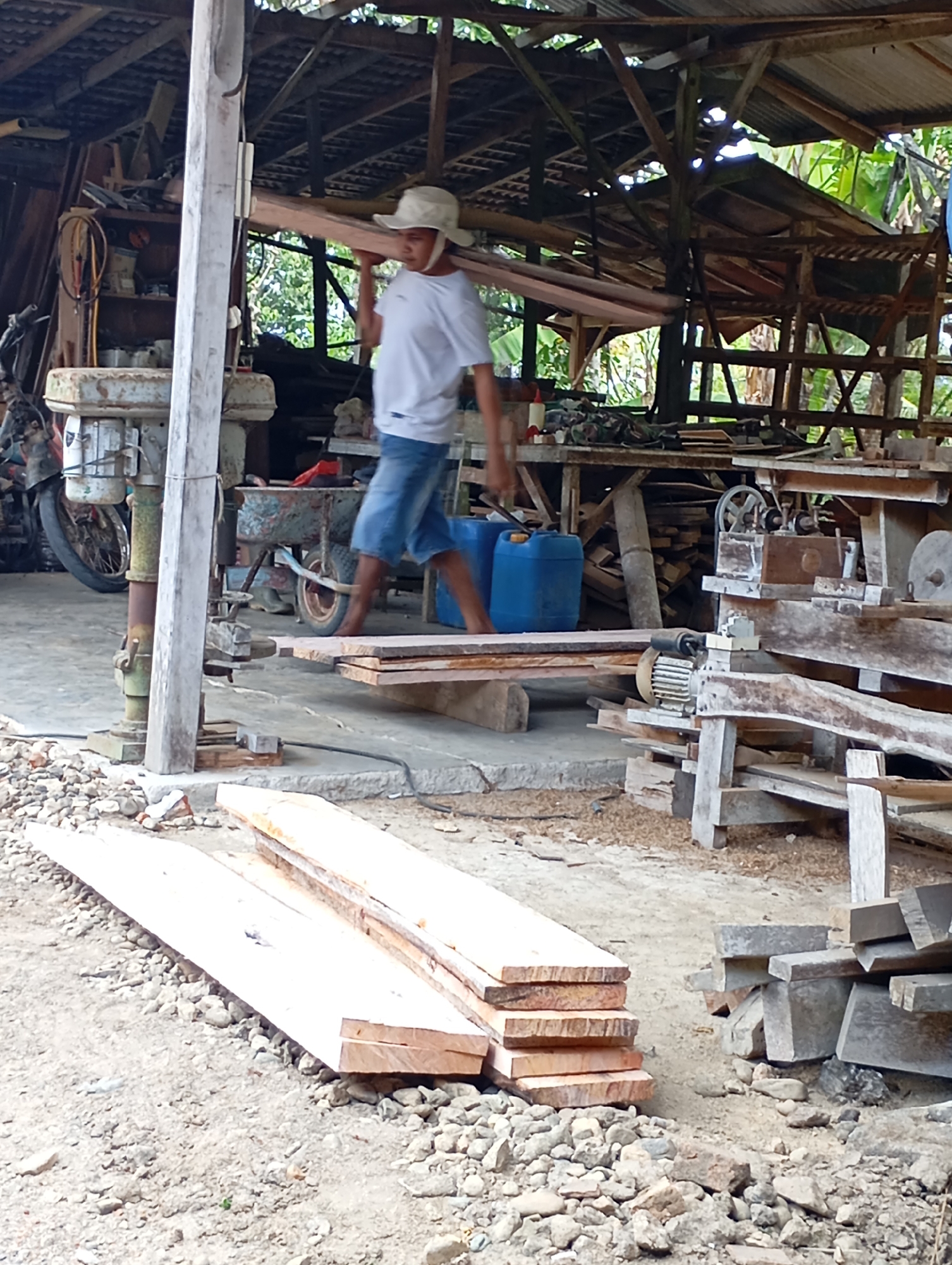 In Ketepung, Indonesia, a man wearing a white t-shirt and denim shorts, with a light-colored hat, carries a long wooden beam across his shoulders within an open-air workshop. The workshop features a corrugated metal roof supported by wooden beams, with various tools and materials visible. To the left, a large industrial drill press stands next to a motorcycle. In the foreground, stacks of lumber are laid out on the ground, composed of light-colored wood planks. The ground is covered in gravel and sawdust. In the background, a wooden workbench is set up with machinery and more wood. Two large blue plastic containers are also present in the middle ground.