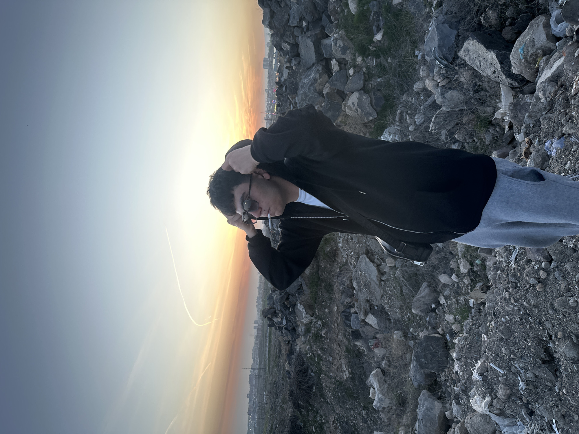 A young man stands outdoors on a rocky, uneven surface at sunset. He wears sunglasses, a black jacket over a white shirt, and gray sweatpants. He has his hands behind his head and a cigarette in his mouth. The background shows a cityscape silhouetted against a colorful sky, with a mixture of urban buildings and industrial structures. The foreground is composed of rough rocks and debris, suggesting a neglected or undeveloped area on the edge of the city.