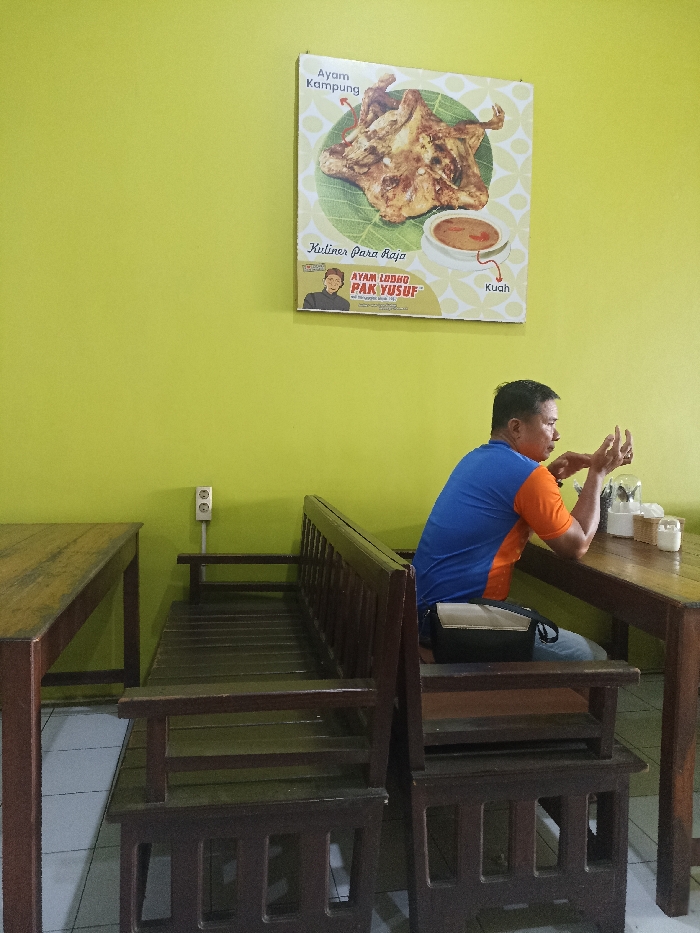 Inside a casual eatery in Tulungagung, Indonesia, a man sits at a wooden table against a bright lime-green wall. He is positioned on the right side of the frame, facing away from the camera but partially turned to his left, gesturing with both hands. He has short dark hair and is wearing a blue and orange short-sleeved polo shirt, along with light-colored pants or jeans. A black and tan shoulder bag rests on the wooden bench next to him.
The eatery features simple, dark wooden furniture, including the table and bench the man occupies, and an identical empty table and bench to his left. On the table, various small white condiment containers and a holder for napkins or cutlery are visible. An electrical outlet with two sockets is mounted on the green wall between the two seating arrangements.
Dominating the upper right portion of the wall is a large rectangular poster advertising a local dish. The poster features an image of a whole roasted chicken, labeled "Ayam Kampung" (free-range chicken), resting on a banana leaf. Beside it is a small bowl of sauce, labeled "Kuah" (sauce/gravy). Below the chicken, text reads "Kuliner Para Raja" (Cuisine for Kings) and features a caricature of a man, with the main dish identified as "AYAM LODHO PAK YUSUF." The man's active hand gestures suggest he might be engaged in a conversation or explaining something. The setting indicates a relaxed, local dining environment.
