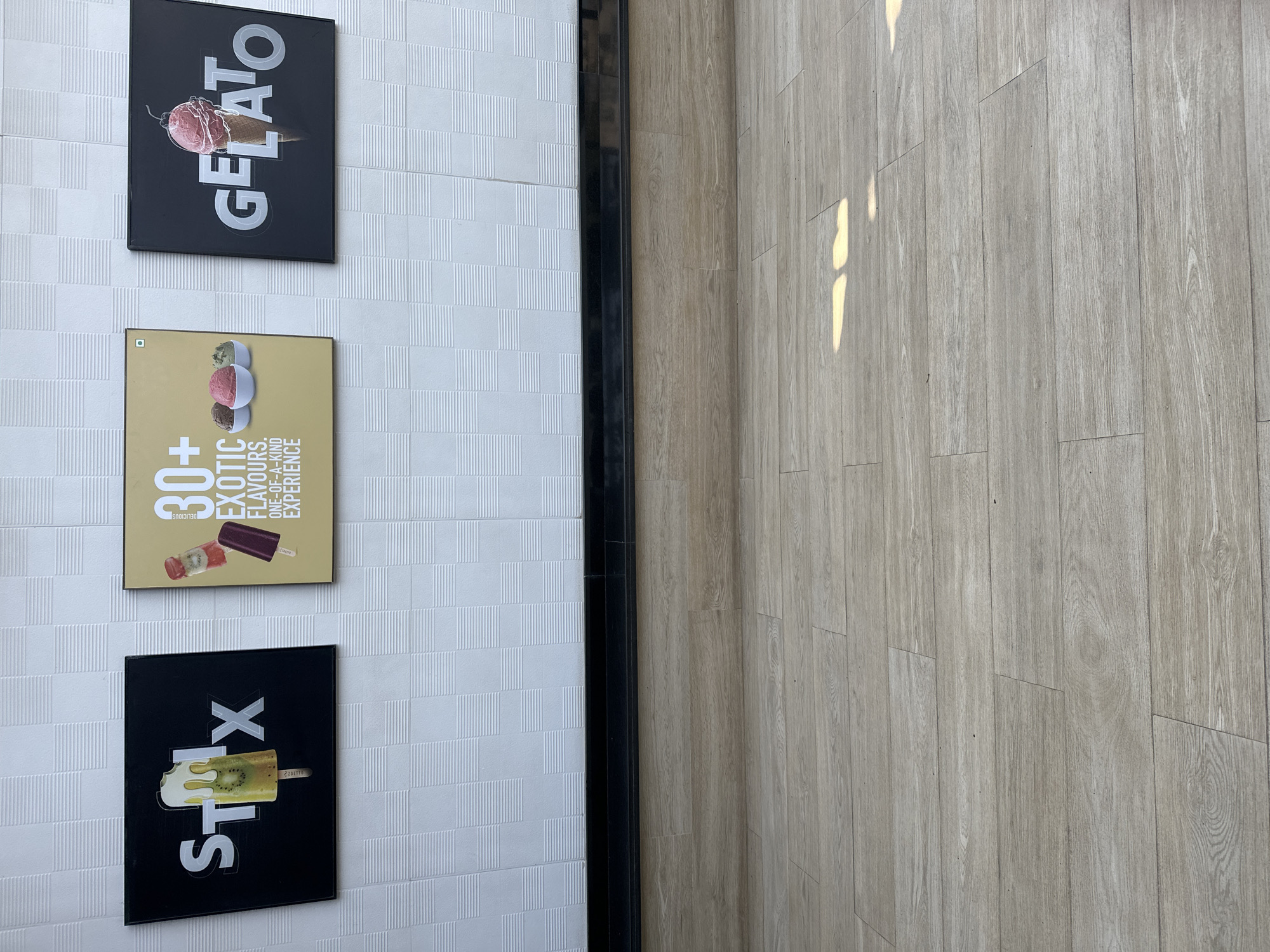 The image shows a section of a wall with three signs and part of a floor. The wall is white with a textured pattern. The signs, from top to bottom, feature:
1. A black sign with the word "GELATO" written vertically in a stylized font, with a picture of a gelato cone.
2. A tan-colored sign with the text "30+ EXOTIC FLAVOURS. ONE-OF-A-KIND EXPERIENCE" and images of frozen treats. A food safety symbol is in the upper left corner.
3. A black sign with the word "STIX" vertically. The "I" is stylized to look like an ice cream bar.
A black vertical frame separates the wall from the floor. The floor is a light brown wood-patterned surface.
Based on the signage related to frozen treats, it is likely that this is a shop or restaurant interior in Mumbai, India.