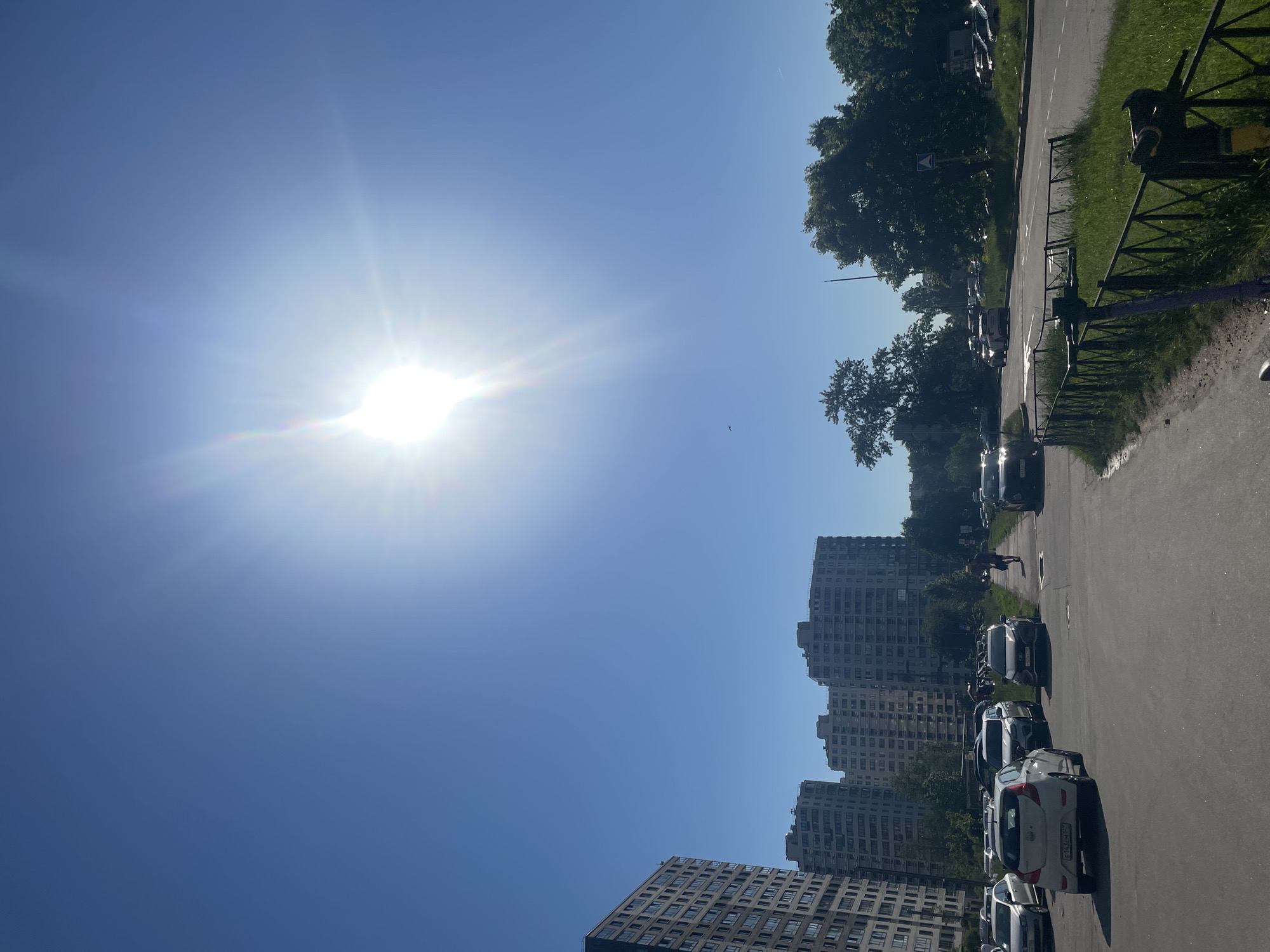 The image displays a sunny street scene dominated by a clear, bright blue sky. The sun, prominent in the upper right corner, casts strong rays and a lens flare across the sky.
On the left side of the frame, several tall, multi-story residential buildings, likely apartment blocks with numerous windows, rise towards the sky. Their architecture appears typical for urban residential areas.
To the right, a paved road stretches into the distance, lined with numerous parked cars. The cars are facing towards the left side of the frame, with some additional vehicles visible further down the street, suggesting ongoing traffic. Green trees and patches of grass border the road, partially obscured by a dark metal fence. A street sign is visible among the trees.
A lone person is seen walking on the pavement next to the road, near the buildings in the mid-ground. A small, dark object, possibly a bird, is discernible high in the clear sky.
The bright, direct sunlight and lack of clouds indicate a clear and likely warm day. The scene captures a moment of everyday life in a residential area. A partially visible license plate on one of the white parked cars reads "C 436 EH," which is consistent with a Russian vehicle registration, confirming the location context.
