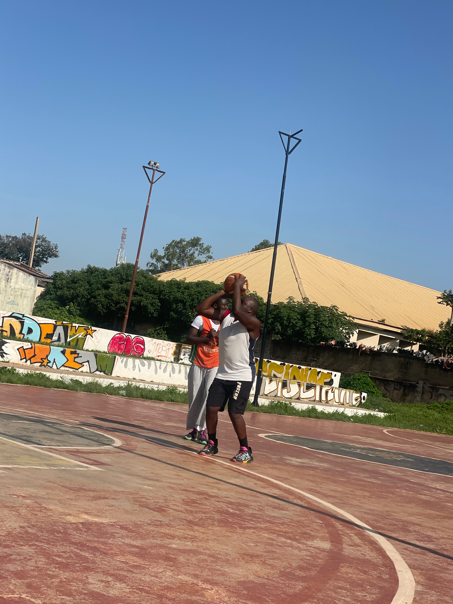 Two young men are playing basketball on an outdoor court with a red and worn surface. The man in the foreground is wearing a white tank top and black shorts, holding a basketball above his head as if preparing to shoot. He has on colorful athletic shoes. The second man, to his left, is wearing an orange basketball jersey and gray pants, and he is also reaching up with his hands.
Behind them, a wall is covered in vibrant graffiti featuring large, stylized lettering in shades of orange, yellow, blue, and green, along with other abstract designs. Beyond the wall, lush green trees form a dense backdrop. In the distance, a building with a yellow, corrugated metal roof is visible. The sky is clear and bright blue. Two tall, dark poles with angular tops stand on either side of the court, likely for lighting or other purposes. One of these poles has a small triangular structure with what appears to be lights at the top. The scene appears to be set in a public space, possibly a park or community center, under the bright Nigerian sun. The overall impression is one of casual athletic activity in an urban environment.