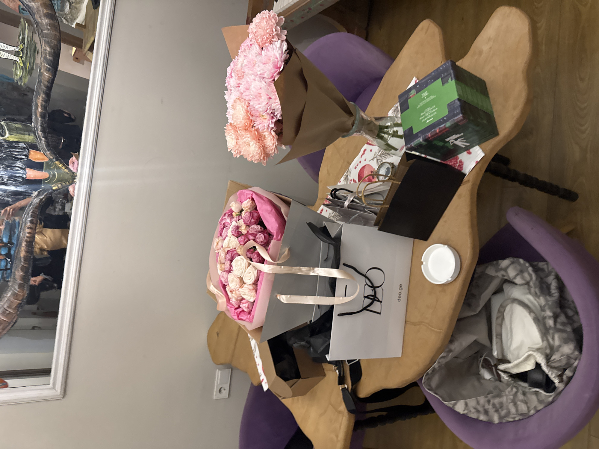 The image depicts a table laden with gifts and flowers in an interior setting. Two bouquets are visible: one wrapped in brown paper with pink and orange flowers, and another in pink paper with pink and white flowers. A green and red box, a black rectangular object, and a white ashtray are also on the table. Shopping bags with the "deo.ge" logo are present, along with eyeglasses. The table has an unusual, organic shape. Three purple chairs surround the table. A framed piece of artwork hangs on the wall.
Given the presence of the "deo.ge" logo, a Georgian e-commerce platform, and the overall setting, it is likely the location is in the city of Tbilisi, Georgia.