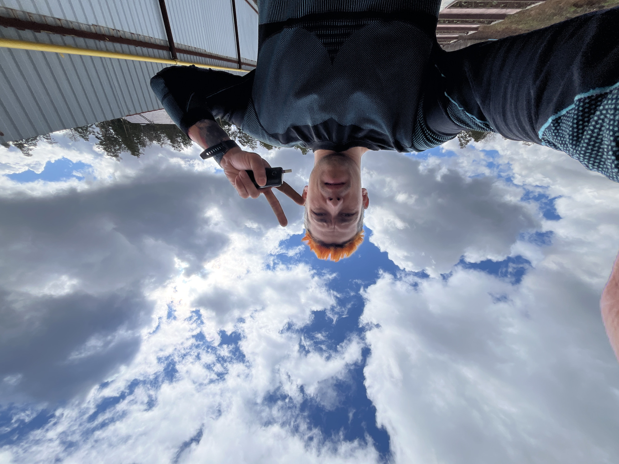 The image is a unique, upside-down selfie, captured from a low-angle perspective looking upwards. The primary subject is a young man with distinctive short, reddish-orange hair, positioned centrally in the frame. He is wearing a dark, long-sleeved athletic shirt, possibly black or dark grey, featuring subtle patterned textures. A dark wristband or watch is visible on his left wrist. The man is holding up his left hand, forming a "V" (peace) sign with his index and middle fingers, while gripping a small, black electronic device, possibly a vape pen, between his thumb and index finger. He gazes directly at the camera with a neutral to slightly smiling expression.
The background is dominated by a dynamic partly cloudy sky, displaying a mix of bright white cumulus clouds and some darker grey clouds, interspersed with patches of clear blue sky. In the upper portion of the inverted image (which would be below when oriented correctly), a light grey corrugated metal roof or awning is visible, with a prominent yellow pipe running horizontally underneath it. Beyond this structure, in the distance, a line of silhouetted evergreen trees can be discerned. The bright, even lighting indicates it is daytime with good visibility. There is no visible text in the image. The setting suggests an outdoor location, potentially an open area or park, with the provided context being Chaikovsky, Russia.