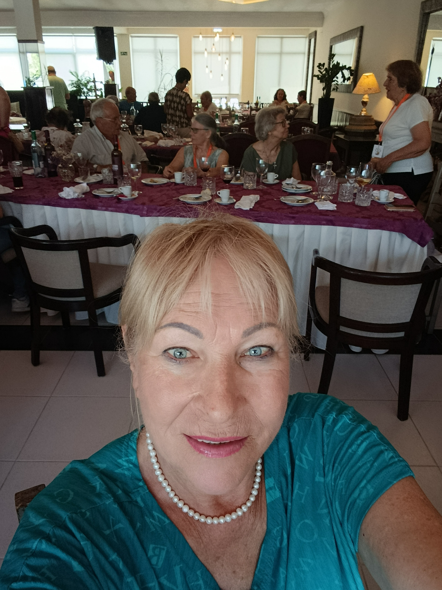 A group of people are seated at a long dining table in what appears to be a banquet hall, likely in Braga, Portugal, given the context. The foreground is dominated by a selfie of a blonde woman wearing a teal top and a pearl necklace. Behind her, the dining table is draped with a purple cloth, laden with place settings, glasses, and bottles. Several individuals are visible seated at the table, engaged in conversation or eating. In the background, large windows with white blinds allow natural light to filter in, and mirrors are mounted on the walls. The overall ambiance suggests a formal gathering or event.