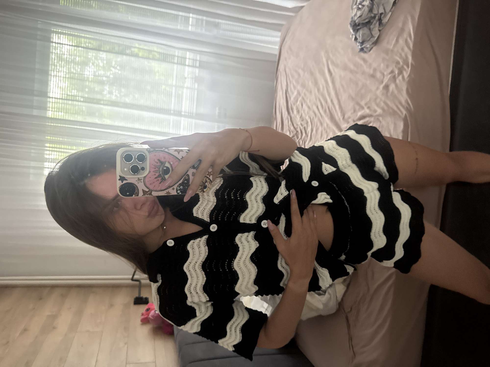 The image, rotated 90 degrees clockwise, depicts a young woman taking a mirror selfie in what appears to be a bedroom during the daytime.
The primary subject is the woman, whose head and upper body are largely visible. She has long, dark hair, which partially covers her face, revealing only her mouth with lips slightly pursed. She is holding a smartphone in her right hand, positioned to capture her reflection. A thin red string bracelet is visible on her right wrist. Her nails are manicured with light-colored polish.
She is wearing a stylish two-piece outfit consisting of a short-sleeved, collared top and matching shorts. Both pieces feature a distinctive black and white wavy striped pattern, possibly knitted or crocheted, with white buttons running down the front of the top. A white undergarment or top peeks out from beneath her patterned shirt.
The smartphone she holds has multiple camera lenses (three are discernible) and is encased in a light pink phone case adorned with a large "evil eye" like design, stars, moons, and other celestial patterns. The case also features glittery or rhinestone embellishments around the camera module.
The background, reflected in the mirror, shows an indoor setting. A large window with white horizontal blinds is prominent, letting in ample natural light. Outside the window, green foliage from trees or bushes is visible, suggesting a clear day. To the right, a bed with light pink or tan-colored bedding, possibly satin or silk, occupies a significant portion of the reflection. Some crumpled fabric, perhaps clothing or a throw, rests on the bed. Below the window, a light-colored wooden floor is visible, and a small pink object, possibly a toy or cushion, can be seen on the floor near the bottom left of the frame. No discernible text is visible in the image.