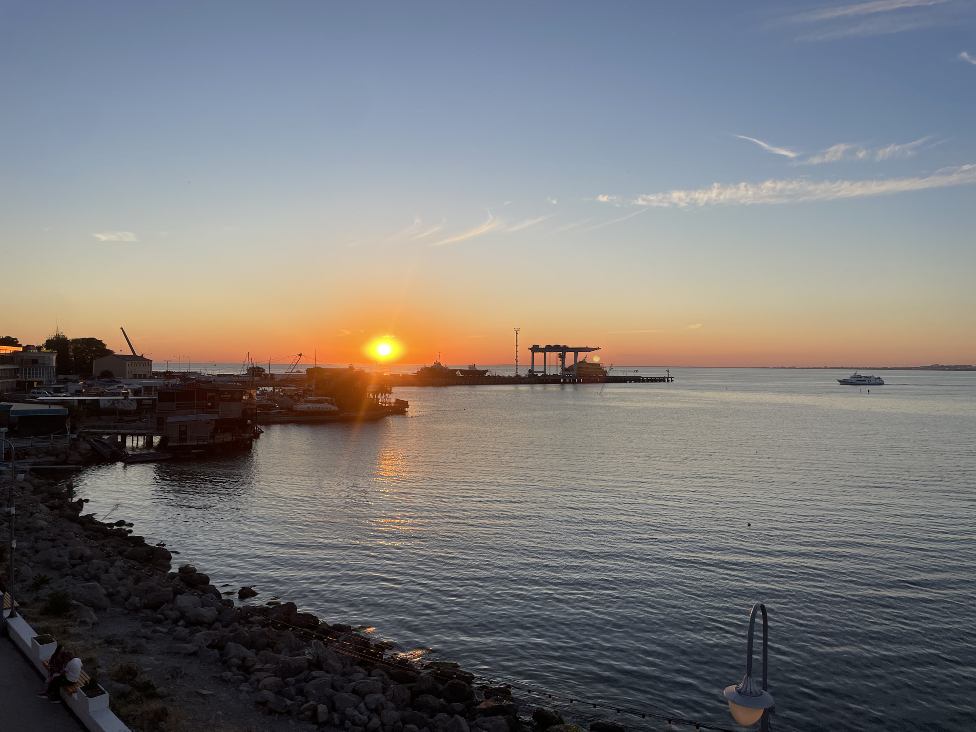 This is a horizontal, eye-level, wide shot of a harbor at sunset in an unknown city. The sun is setting on the left side of the frame, casting a warm orange glow across the sky and water. The sky is clear with a few wispy clouds. The water is calm and reflects the colors of the sunset.
In the foreground, there is a rocky shoreline with a paved path along the top. A few people are sitting on a bench on the path. The water laps gently against the rocks.
In the middle ground, there are several boats docked in the harbor. Some of the boats appear to be fishing boats, while others are larger vessels. There are also some buildings along the waterfront, including what appears to be a restaurant or bar.
In the background, there is a large industrial area with cranes and other equipment. A ferry is visible in the distance, sailing across the water. The overall scene is peaceful and serene, with the warm colors of the sunset creating a beautiful and tranquil atmosphere.