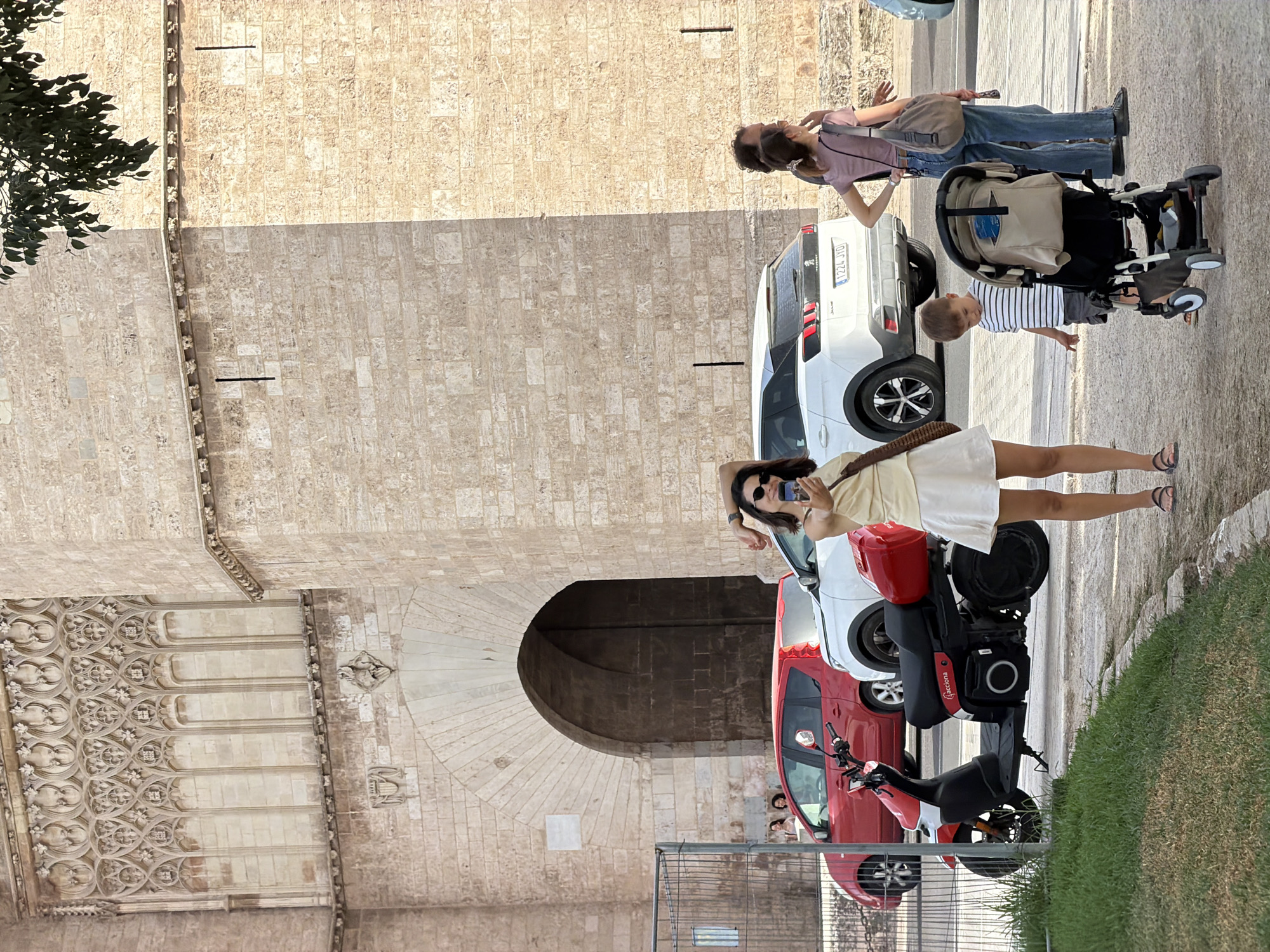 In the city of Unknown, Unknown, a woman is seen taking a selfie in front of a stone building with an arched entrance. She is wearing a yellow top and a white skirt. To her left, a red scooter is parked. Behind the scooter is a red car, and next to it is a white SUV. A woman and a young boy are standing near the SUV, with the boy looking at a stroller. The background features a large stone wall with intricate carvings and a tree branch with leaves hanging from the top right corner of the image. The lighting suggests it is daytime.
