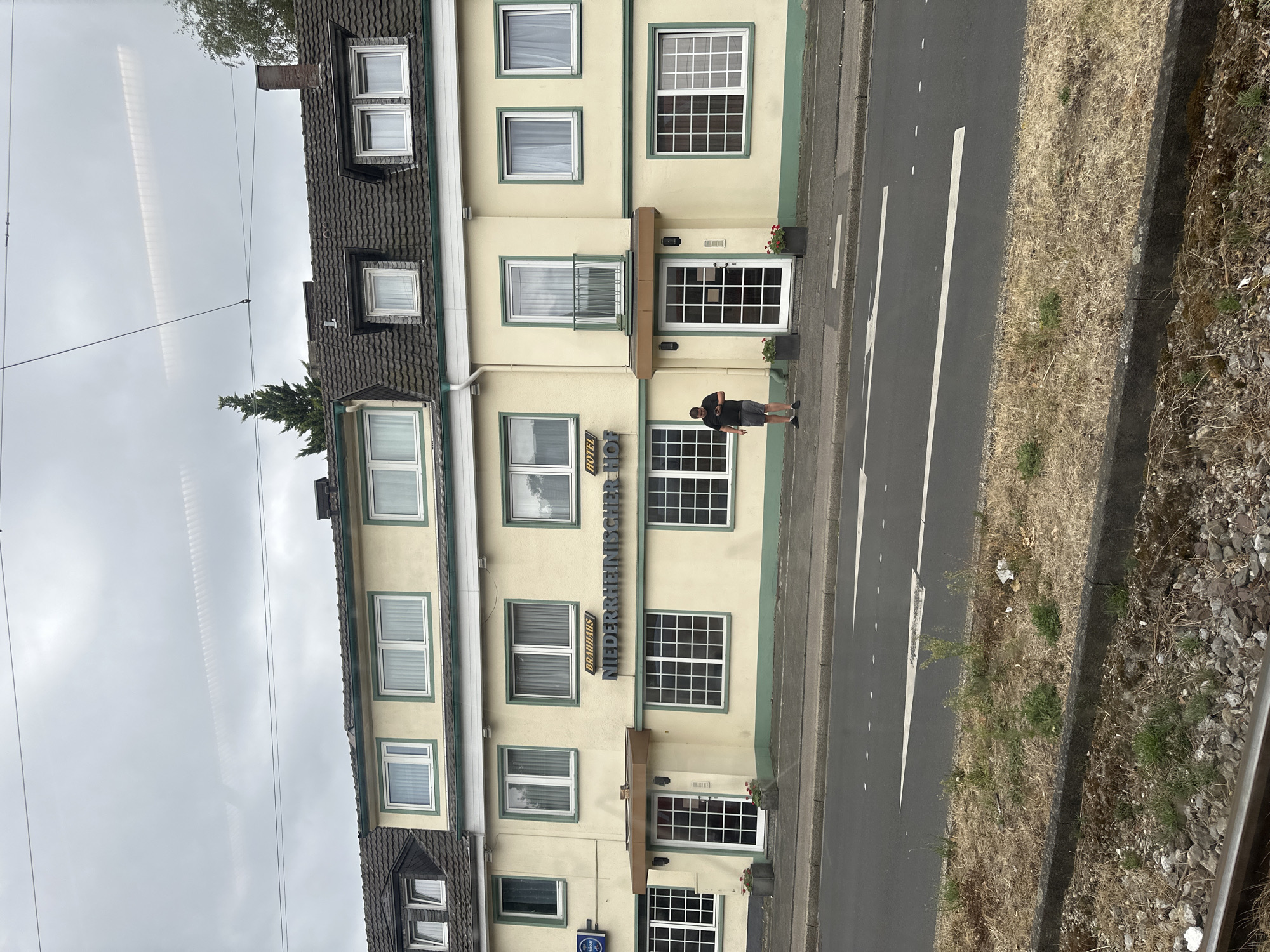 The image shows the exterior of the "NIEDERRHEINISCHER HOF" hotel in Krefeld, Germany. The hotel is a multi-story building with a pale yellow facade, green trim around the windows, and dark tiled roofs. Several windows are visible, some with white curtains drawn. On the ground floor, there is a central entrance with a dark door and a brown awning. A man in a black t-shirt and grey shorts is standing on the sidewalk to the left of the entrance, looking at his phone. To the left of the hotel entrance, a blue sign with white lettering is partially visible. In front of the hotel, there is a sidewalk and a road with lane markings. A grassy area with dry vegetation and some gravel is located between the sidewalk and what appears to be a railway track in the foreground. Overhead, power lines are visible against a cloudy sky.